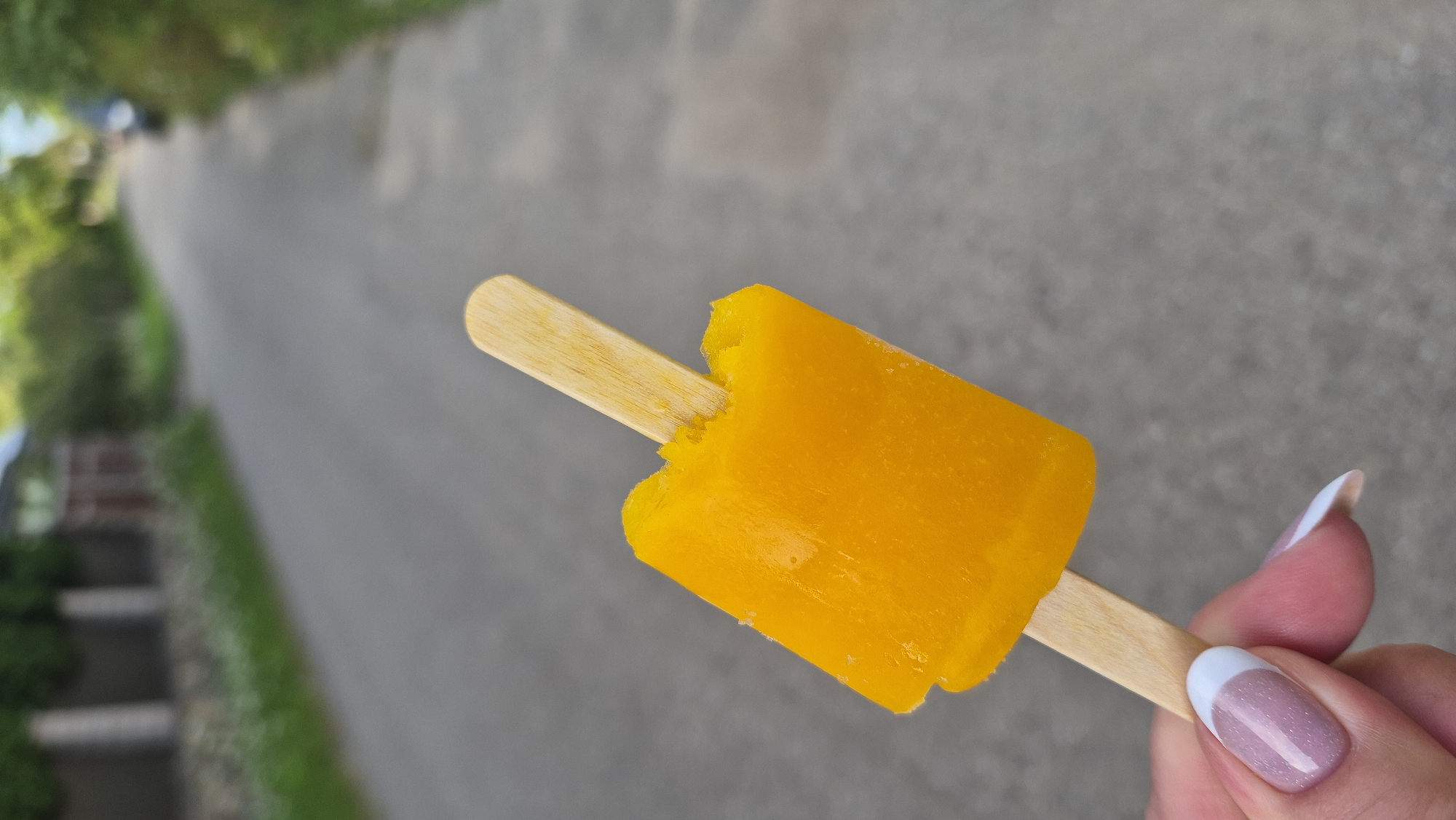 An orange popsicle with a bite taken out of it is held by a person's hand in the foreground. The popsicle is on a wooden stick. The background is blurred and shows a paved road leading into the distance, flanked by greenery and some buildings. The setting appears to be a street in Krauja, Latvia. The hand holding the popsicle has well-maintained fingernails with French manicures. The lighting suggests it is daytime.