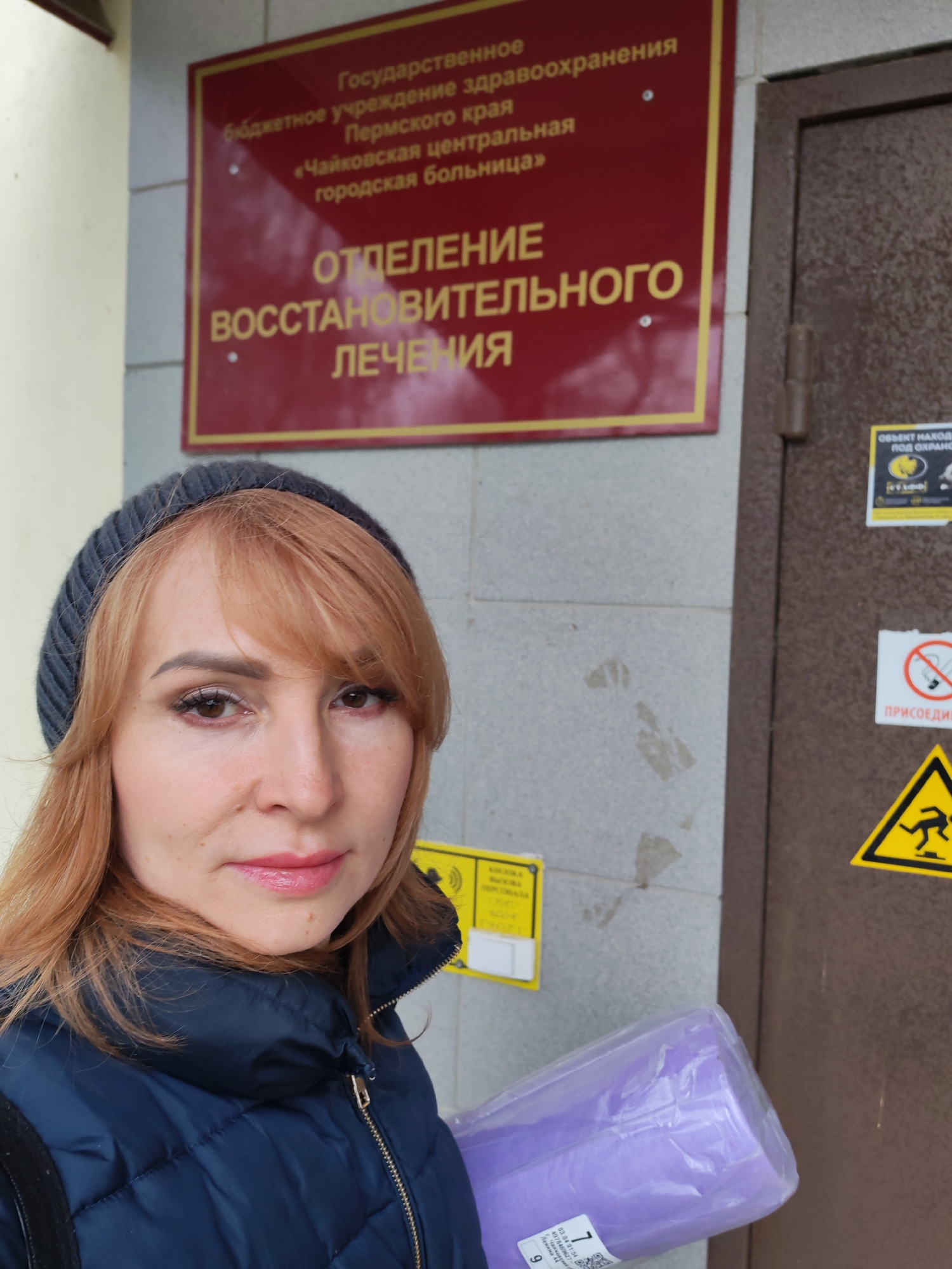 A woman wearing a gray beanie and a blue quilted jacket is posing for a selfie in front of a building in the city of Chaikovsky, Russia. The sign above her reads "Department of Rehabilitation Treatment" of the Chaikovsky Central City Hospital. She is holding a package wrapped in purple plastic. A yellow sign is partially visible to the left. To the right is a door with several warning stickers.