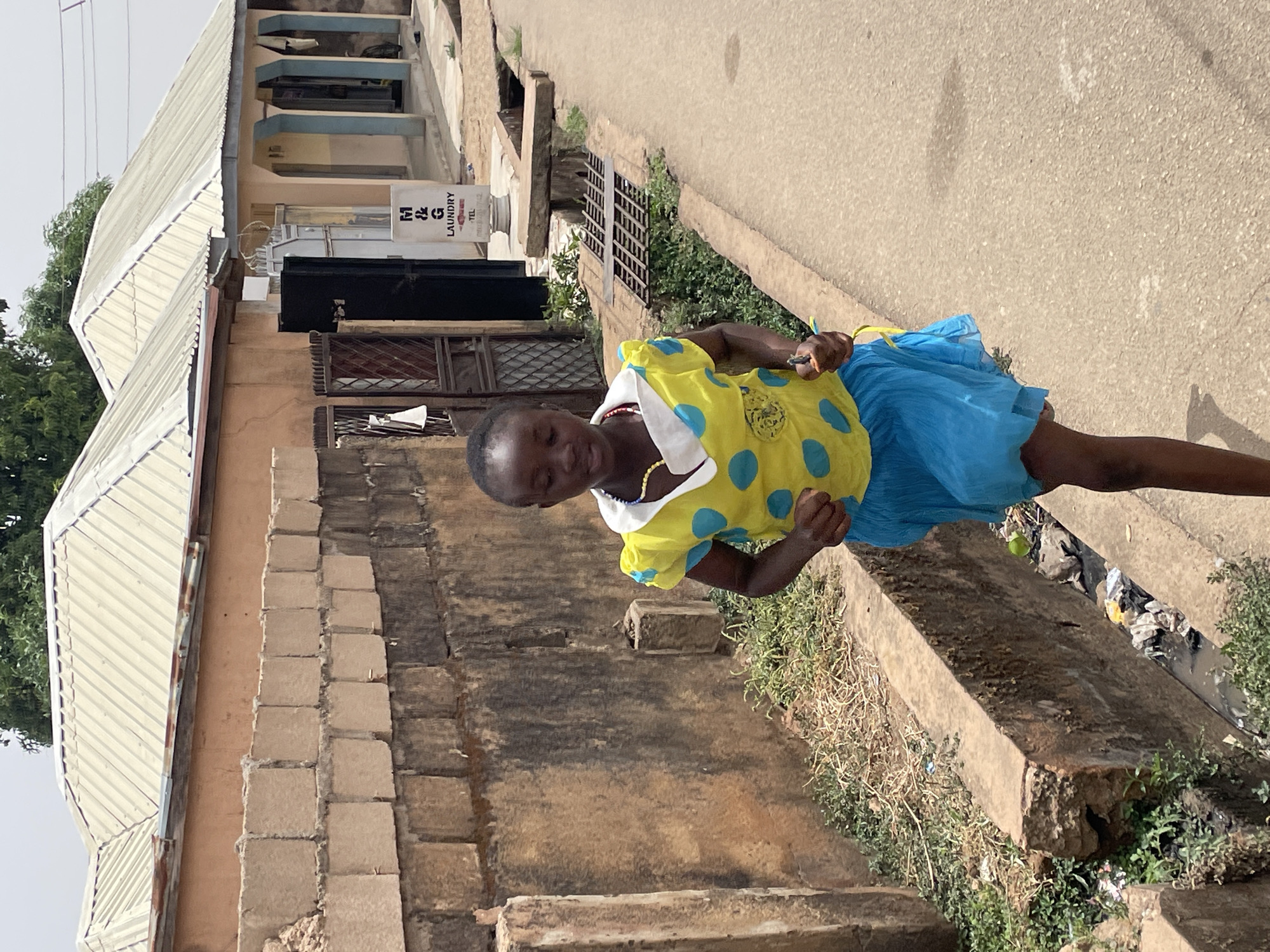 A girl wearing a yellow, blue-polka-dot blouse and blue skirt stands outdoors next to a dilapidated building with brick and concrete construction. The building has a corrugated metal roof. She is facing forward, with her arms bent and hands clenched near her chest. The location is Jos, Nigeria. A sign for M & G Laundry is visible on the building. The street is visible with a cement walkway. Debris and vegetation are along the walkway.