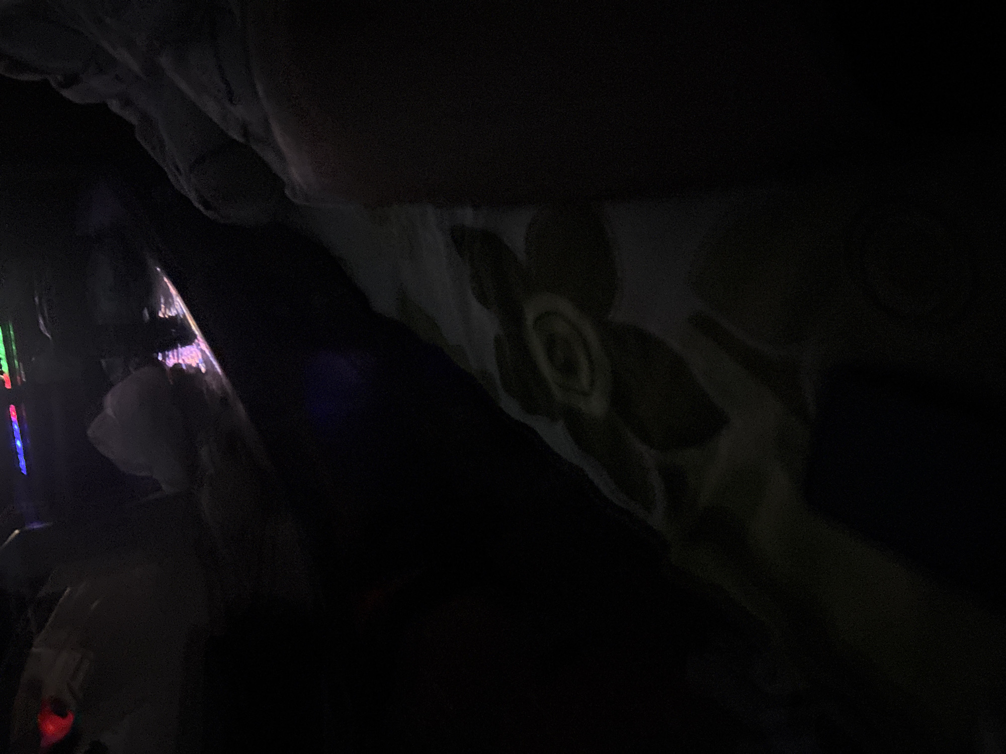The image is dark and underexposed, obscuring many details. A patterned surface with a large floral motif is visible on the right side of the image. On the left, a blurred object with illuminated elements, possibly a device with a screen or lights, is present. The overall environment seems to be dimly lit, potentially indoors. There is no discernible information to identify this location being in the city of Yerevan, Armenia.