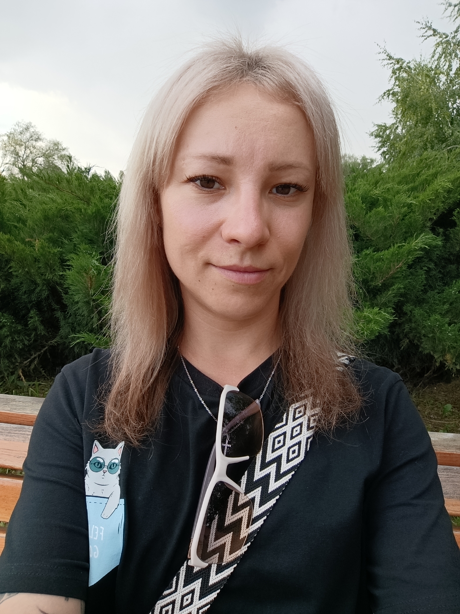 A young woman with shoulder-length blonde hair and brown eyes takes a selfie outdoors. She is wearing a black t-shirt with a light blue pocket print of a white cat wearing sunglasses. A white necklace with a small cross pendant is visible, along with white-rimmed sunglasses hanging from her shirt. She is sitting on a wooden bench, with green trees and bushes in the background. The sky is overcast, suggesting it might be daytime, possibly in the late afternoon. The overall mood is relaxed and casual. The location context is Novokuybyshevsk, Russia.