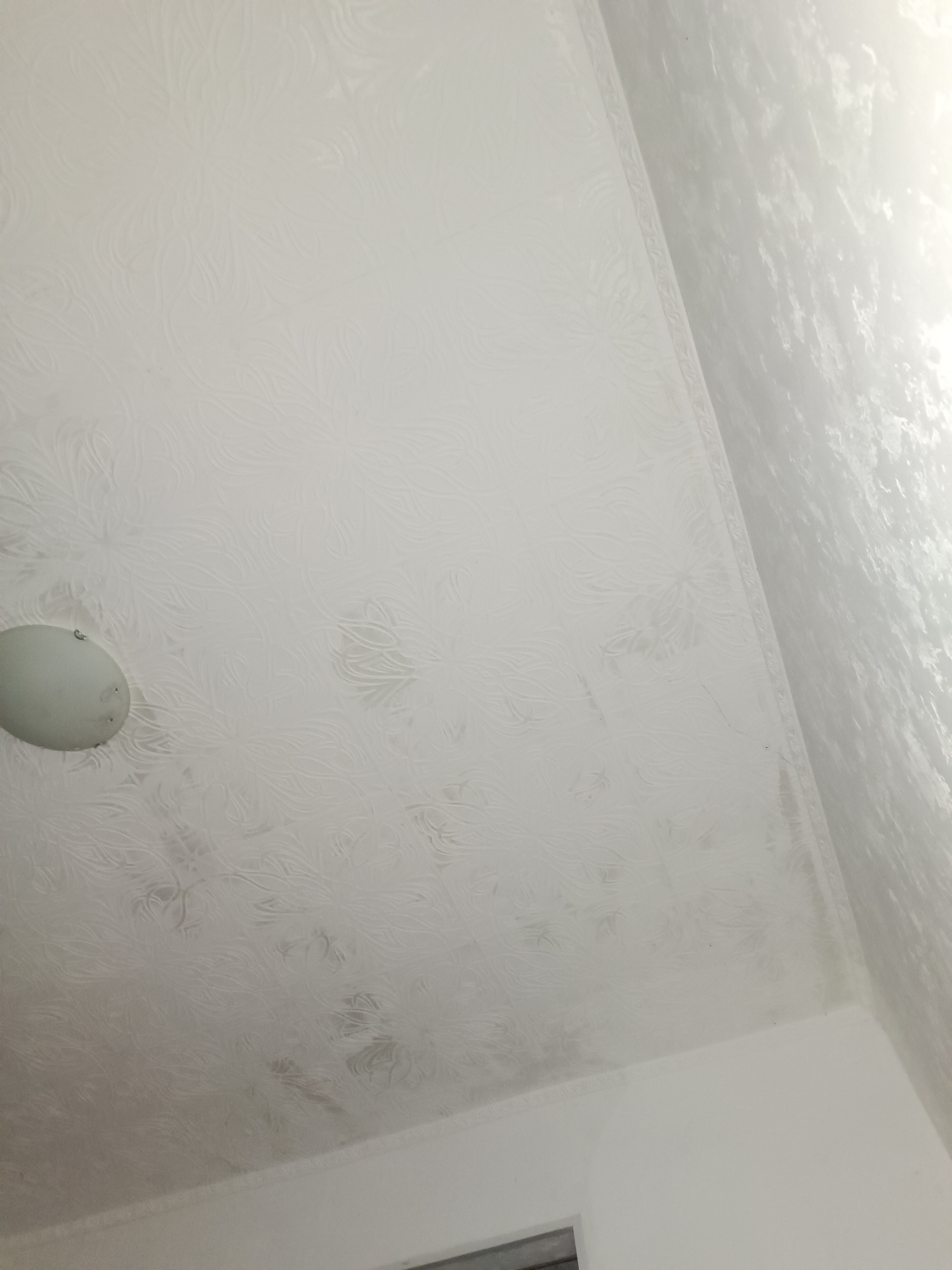 The image shows a close-up view of a textured white ceiling, likely in a residential interior in Saky, Ukraine. A circular light fixture is visible on the left side of the ceiling. The ceiling has a repeating embossed pattern resembling floral or organic shapes. The texture appears to be a common type of ceiling covering, possibly made of polystyrene or vinyl. The lighting suggests it is daytime, with light coming from an unseen source to the right, creating a bright area on the right portion of the ceiling. The overall impression is of an ordinary, well-lit interior space.