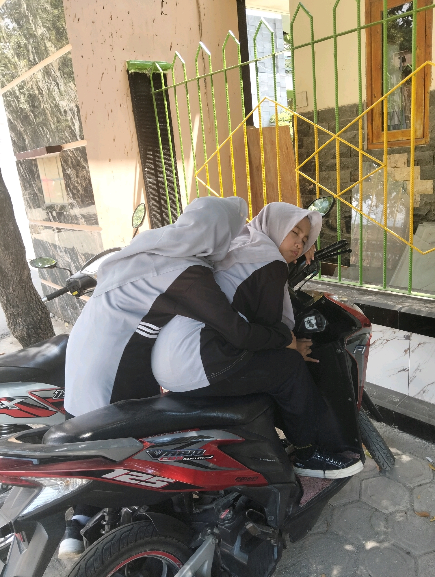 Two young females, wearing hijabs and modest clothing, are seated on a red and black scooter. The female in the foreground, wearing a grey hijab and black pants, is resting her head on her arm and appears to be asleep or resting. She is wearing black sneakers. The female behind her, wearing a grey hijab and white top, is holding her. They are positioned in front of a building with a textured facade and a yellow and green metal fence. The scene appears to be outdoors, possibly in a residential area, in Paciran, Indonesia.