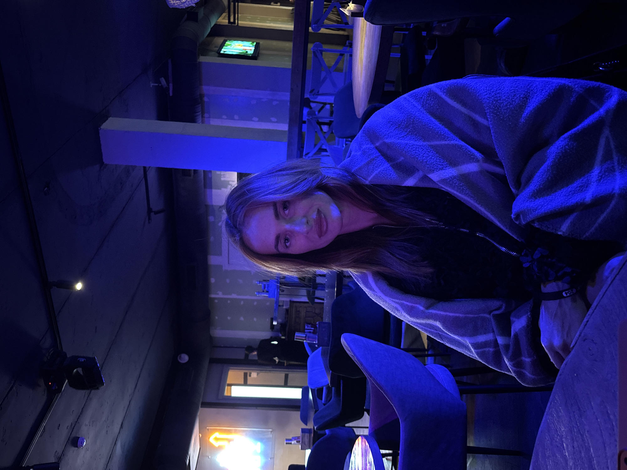The image shows a woman sitting at a table inside what appears to be a bar or restaurant. She is partially covered with a striped blanket or shawl. The lighting is predominantly blue. In the background, there are tables and chairs, and a neon sign with an arrow can be seen through a doorway. A television is mounted on a wall in the background. It is unclear if the location is Krasnoyarsk, Russia.
