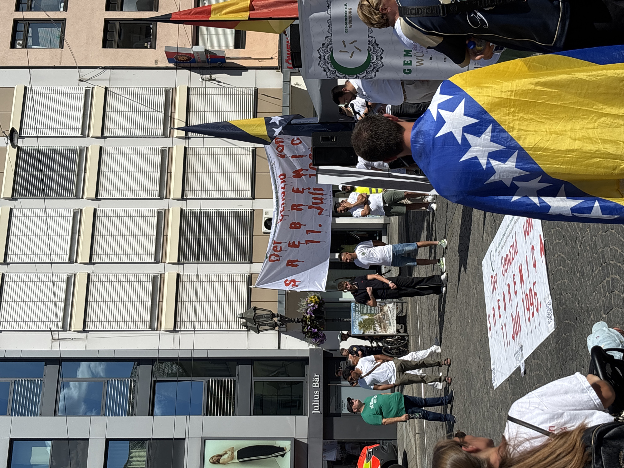The image shows a gathering of people in a city square, likely a protest or memorial event. A large banner hangs in the background with red lettering that appears to be in German and possibly Bosnian, referencing "Srebrenica" and "1. Juli" (July 1st). Several individuals are visible, some wearing casual clothing, others in more formal attire. A man in the foreground is draped in the Bosnian flag. Flags of Germany are also visible. In the background, buildings with modern architecture and awnings can be seen, as well as a street sign indicating "Julius Bär". Another sign on a display board reads "ZUKUNFTSALLEE DERARBEITERWURZELN" and mentions "IGBD-Gemeinde e.V. Würzburg". This suggests the location is Würzburg, Germany. The ground is paved with cobblestones, and several banners and flags are displayed. People are standing around, some appearing to listen to a speaker or observe the events. The scene is illuminated by daylight, indicating it is likely daytime.