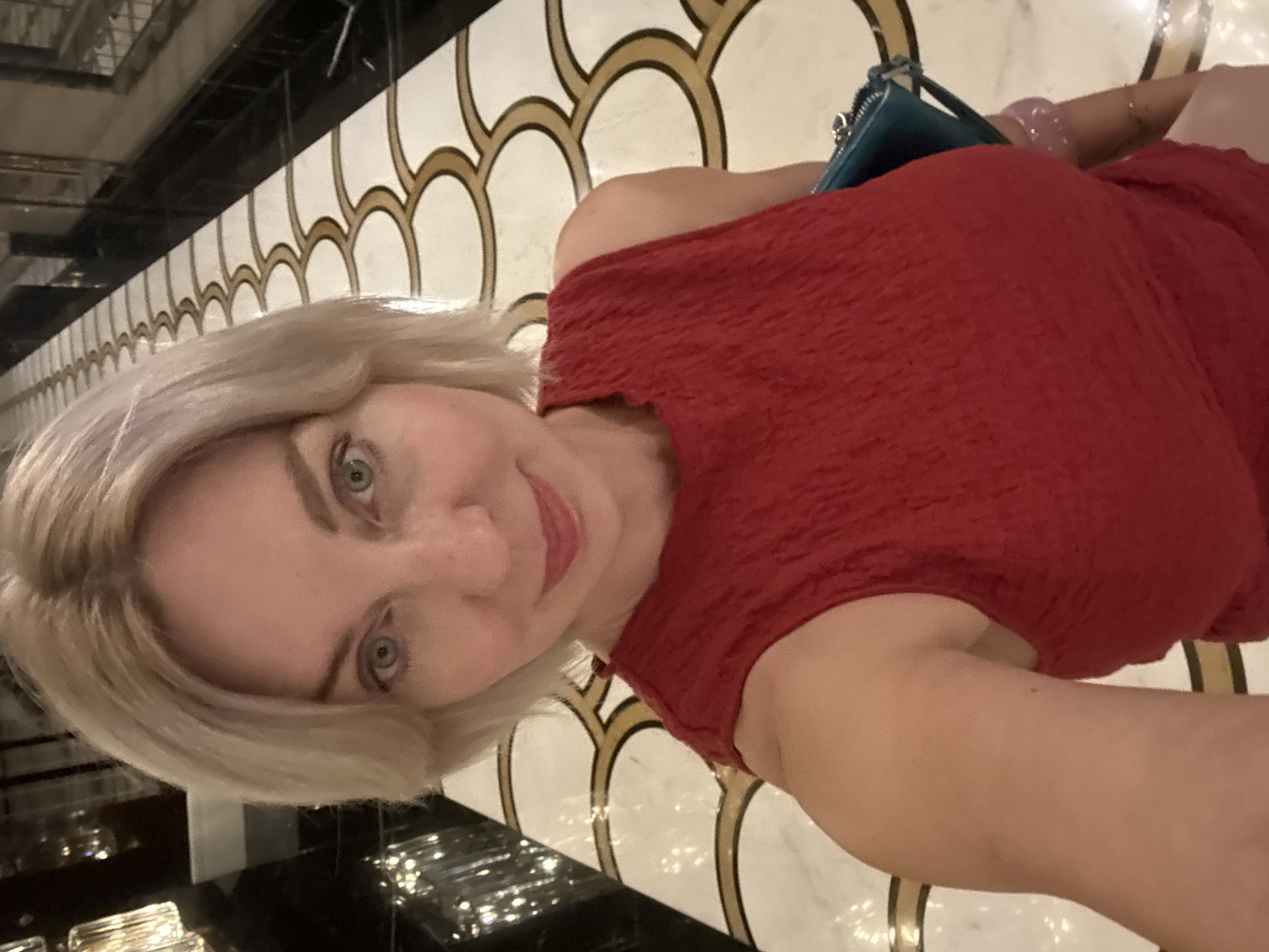 A blonde woman with blue eyes and fair skin, wearing a red textured sleeveless top and holding a teal clutch, stands in a hallway with a patterned marble floor and decorative lighting. The hallway features a repeating scalloped pattern on the floor in white and gold. The woman is looking at the camera and smiling. She is wearing a few bracelets on her left arm. The image appears to be a selfie taken indoors. The specific location is stated as Estosadok, Russia, implying a hotel or resort setting.