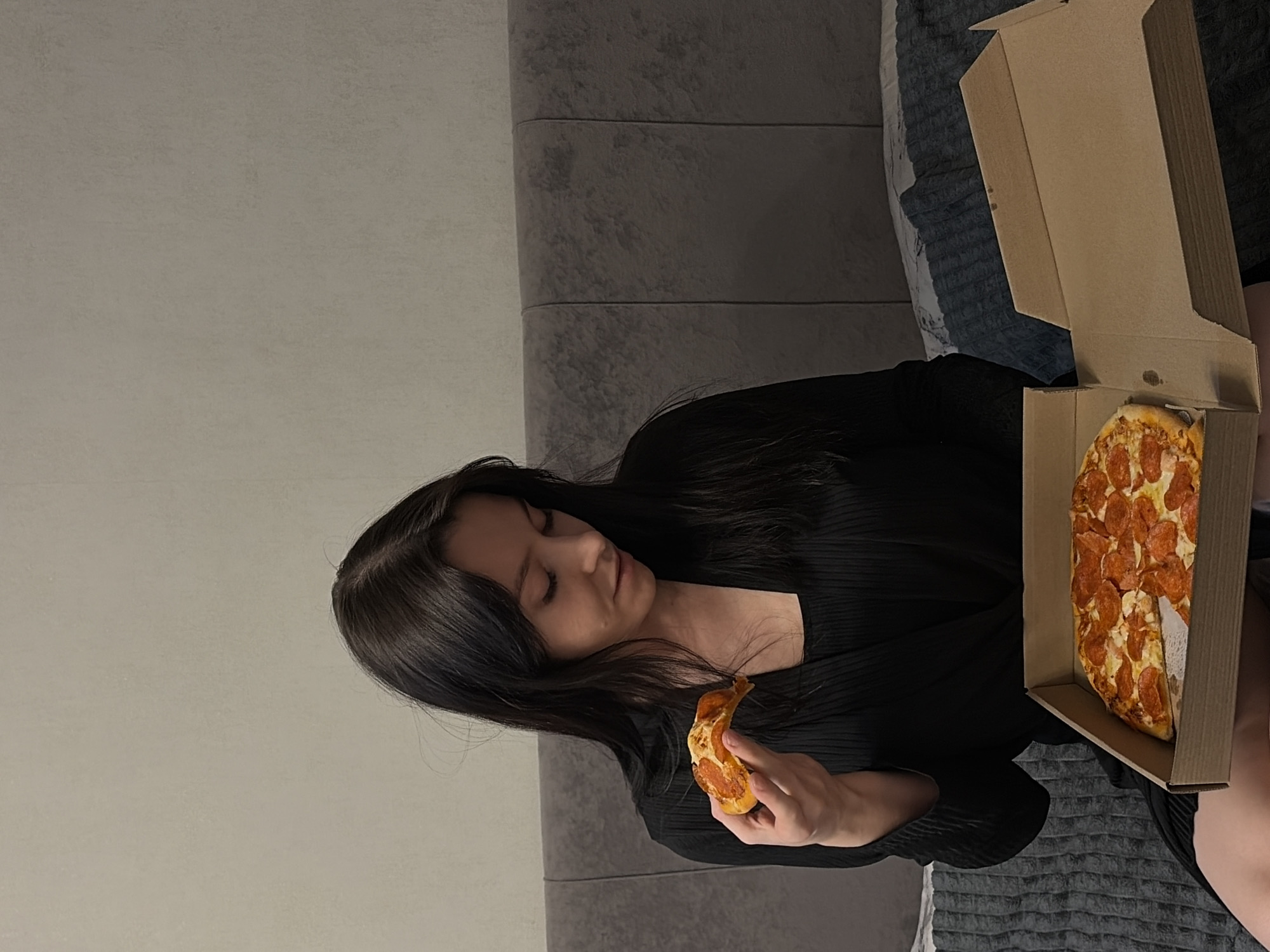 A woman is sitting indoors, possibly on a couch or bed with grey bedding. She is holding a slice of pizza in her left hand, while an open pizza box filled with pepperoni pizza sits beside her. She has long dark hair and is wearing a black top. The background consists of a plain grey wall. The location is not discernible from the image.