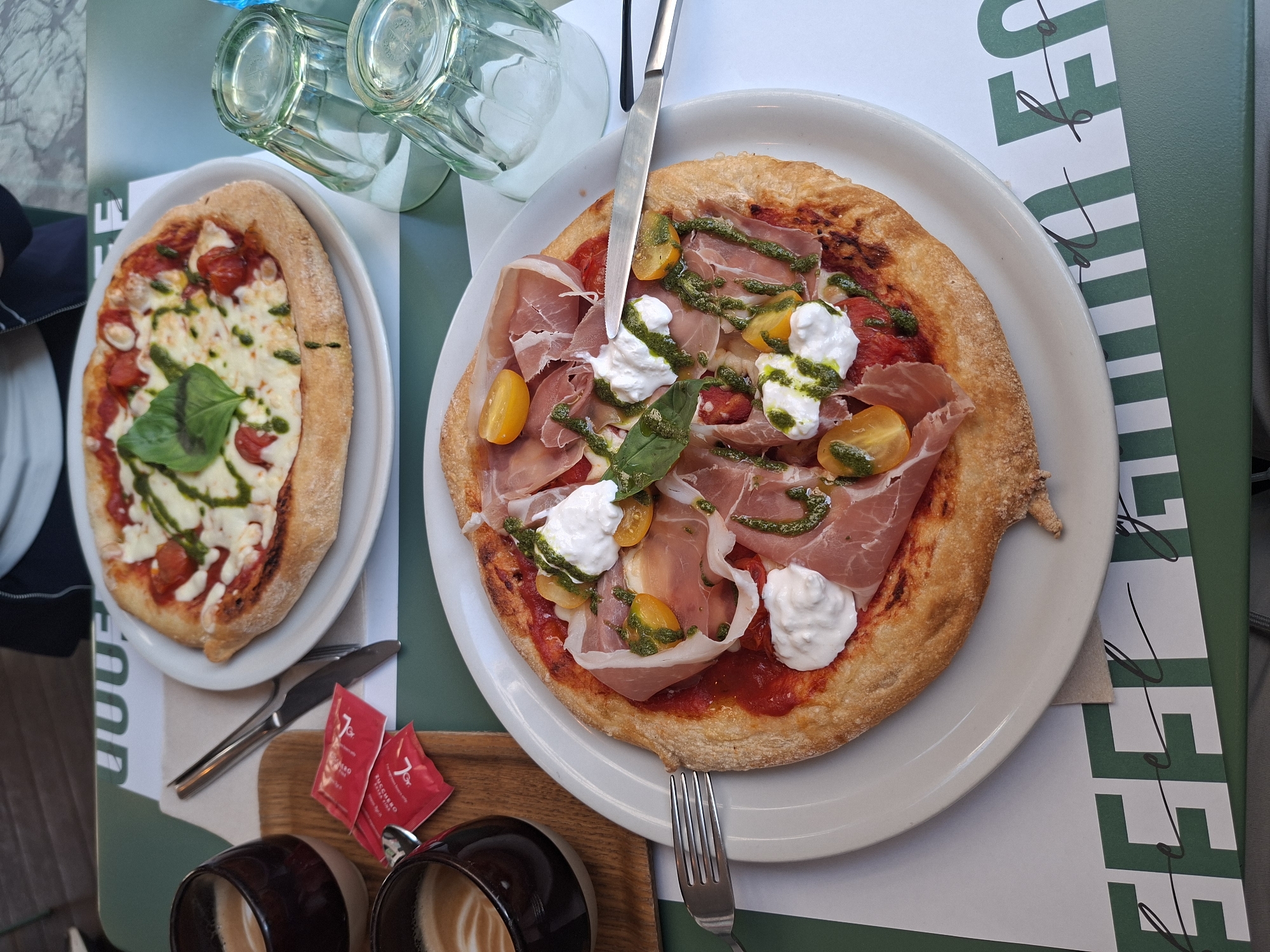 The image shows a table at what appears to be an outdoor restaurant or cafe in Milan, Italy. Two pizzas are presented on white plates. The pizza on the left has a Margherita style with tomato sauce, mozzarella, cherry tomatoes, and a basil leaf. The pizza on the right is topped with tomato sauce, mozzarella, prosciutto, yellow cherry tomatoes, and pesto sauce. Silverware is positioned next to each pizza. Two empty glasses are visible above the pizzas. Below the pizzas are two cups containing coffee drinks with latte art, along with two sugar packets. The table has a green and white design.