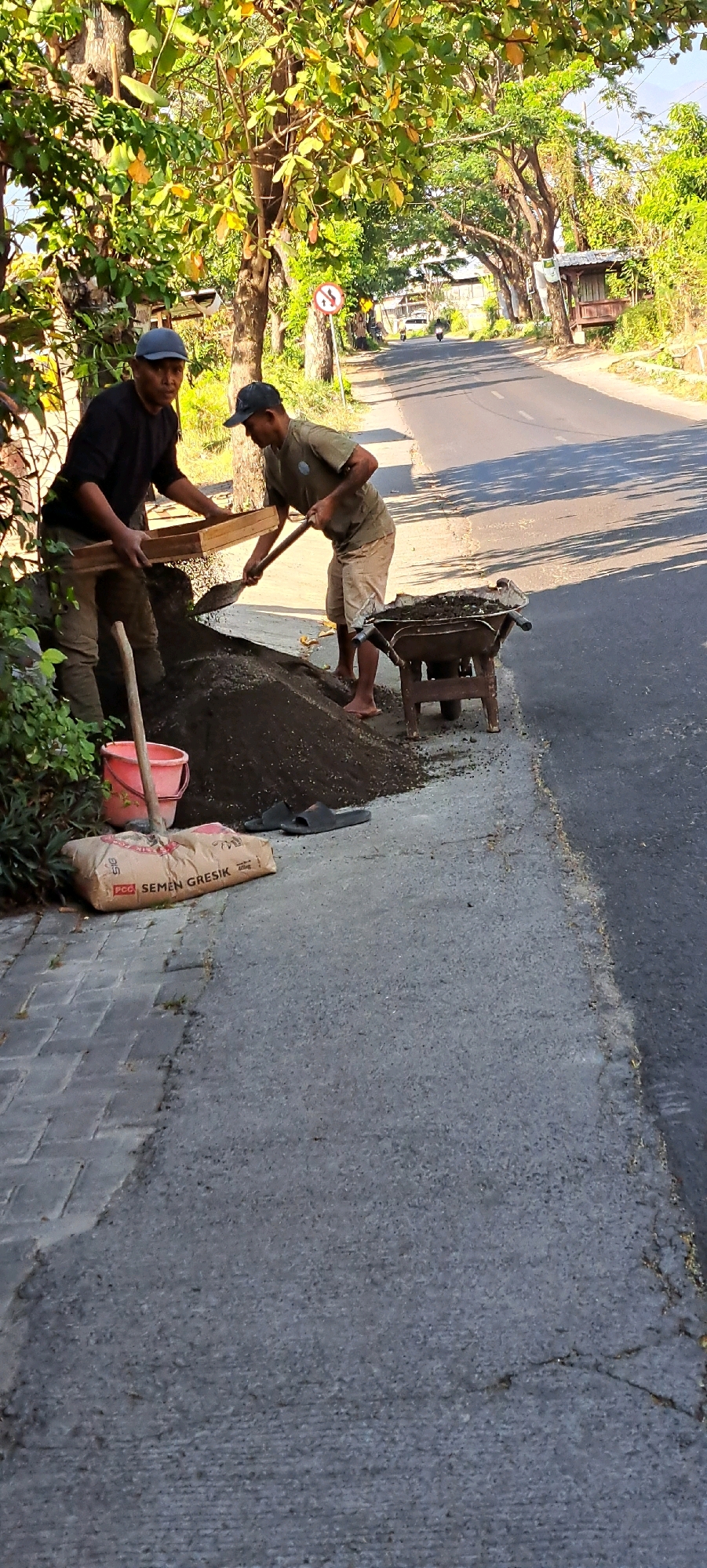 Two individuals are engaged in construction work by a paved road in Sumber Gedang, Indonesia. The person on the left, wearing a black shirt and a blue cap, is holding a sieve over a pile of dirt. The person on the right, in a green t-shirt and shorts, is using a shovel to add dirt to the sieve. A wheelbarrow filled with dirt stands nearby, along with a red bucket and a bag of "SEMEN GRESIK" cement. The scene is set under the shade of trees lining the road, with a residential area visible in the background. The lighting suggests it is daytime, with shadows cast by the trees on the road.
