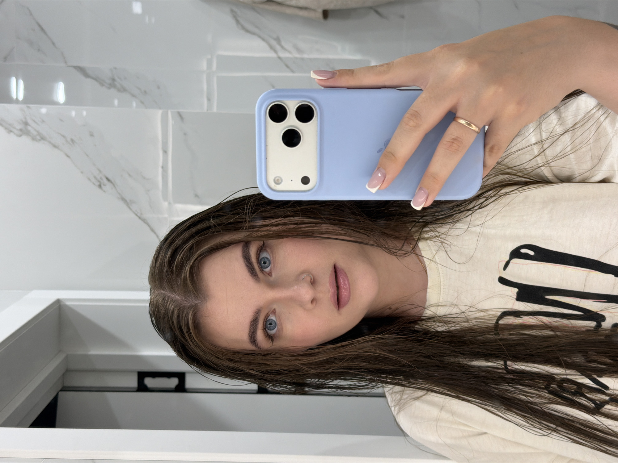 In a brightly lit bathroom with marble-patterned walls, a young woman with long, wet brown hair is taking a selfie in a mirror. She is wearing a light-colored t-shirt with a black graphic design. Her eyes are a striking blue, and she is wearing a nose ring. She is holding a light blue smartphone with a multi-lens camera in her right hand. Her nails are manicured with a French tip design, and she is wearing a thin gold ring on her left ring finger. The background shows a door frame and a portion of a tiled wall. The location is identified as Chaikovsky, Russia.