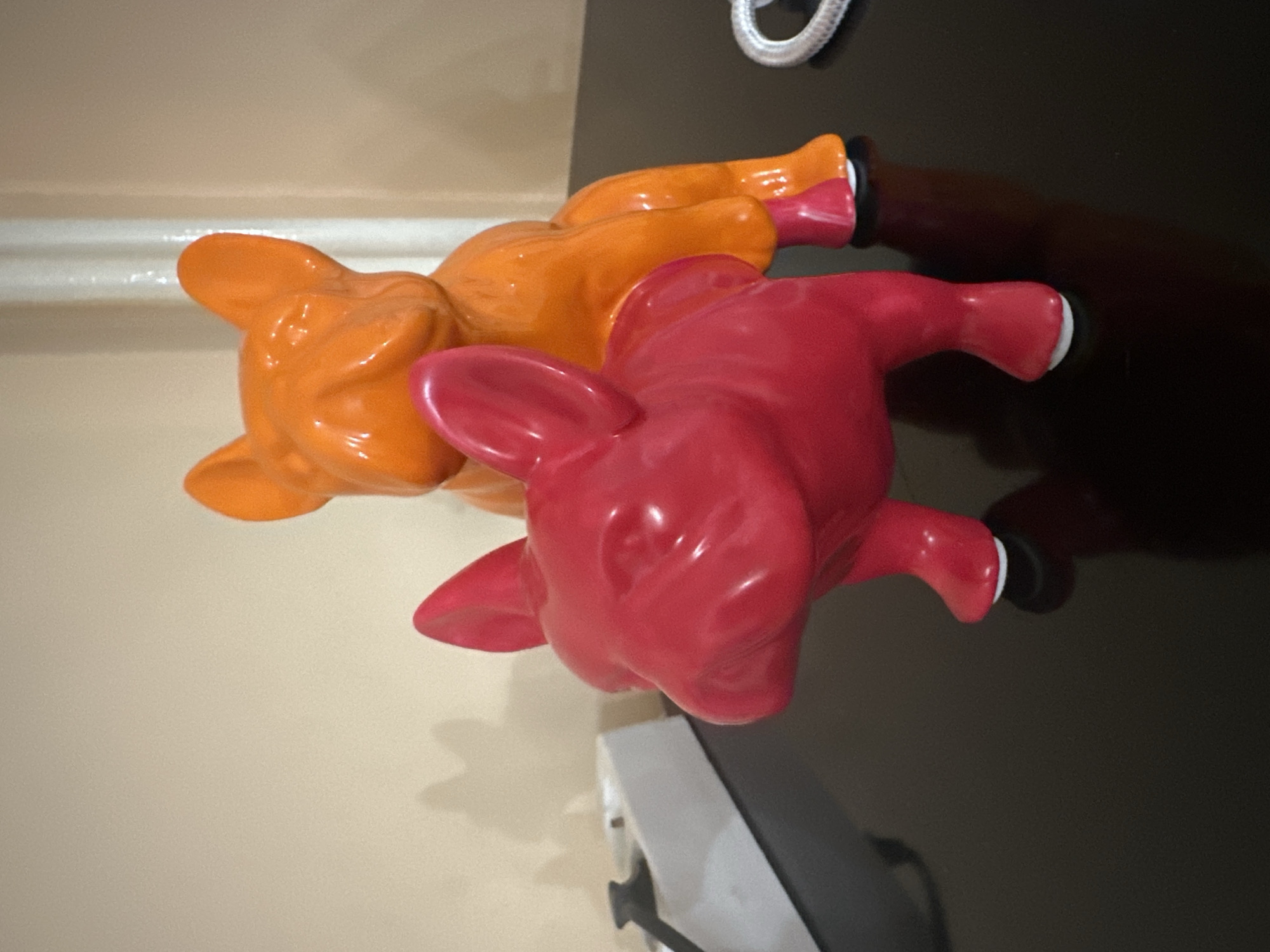 Two ceramic pig figurines, one orange and one red, are positioned on a dark, reflective surface, likely a table or countertop. The orange pig is partially obscured by the red pig, which is in the foreground. The surface also has other miscellaneous objects like an electrical outlet, a power adapter, and a silver cable. The background is a light beige wall.