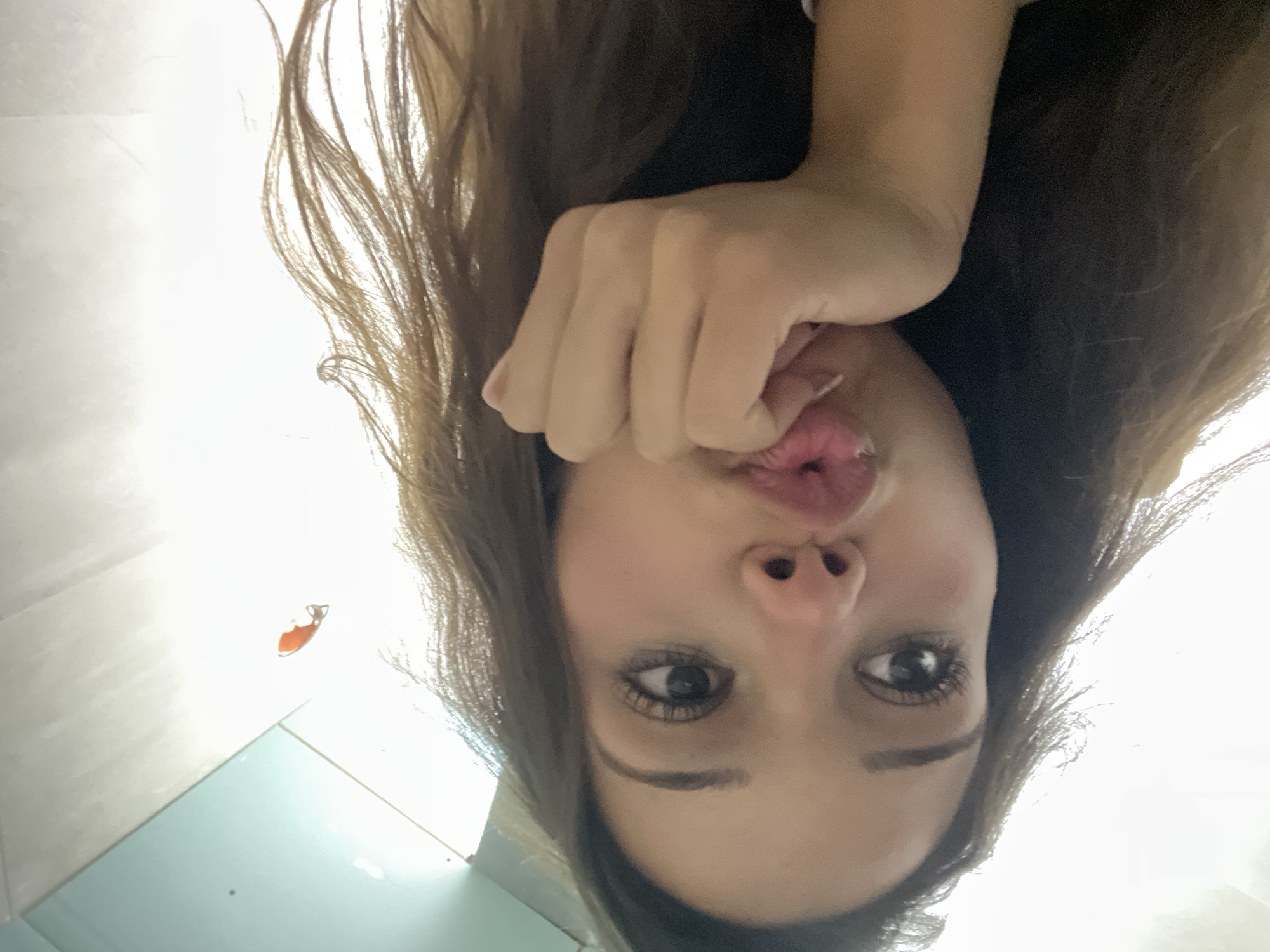 A close-up, low-angle selfie features a young woman with long, brown hair. Her hand rests on her chin, and her lips are puckered. She is looking to her left, and her eyes are wide open. The background is a bright, white wall with a light blue ceiling. There is an orange object, possibly a sticker, on the wall in the upper right corner. There is no indication of Zlatograd, Bulgaria, in the image.