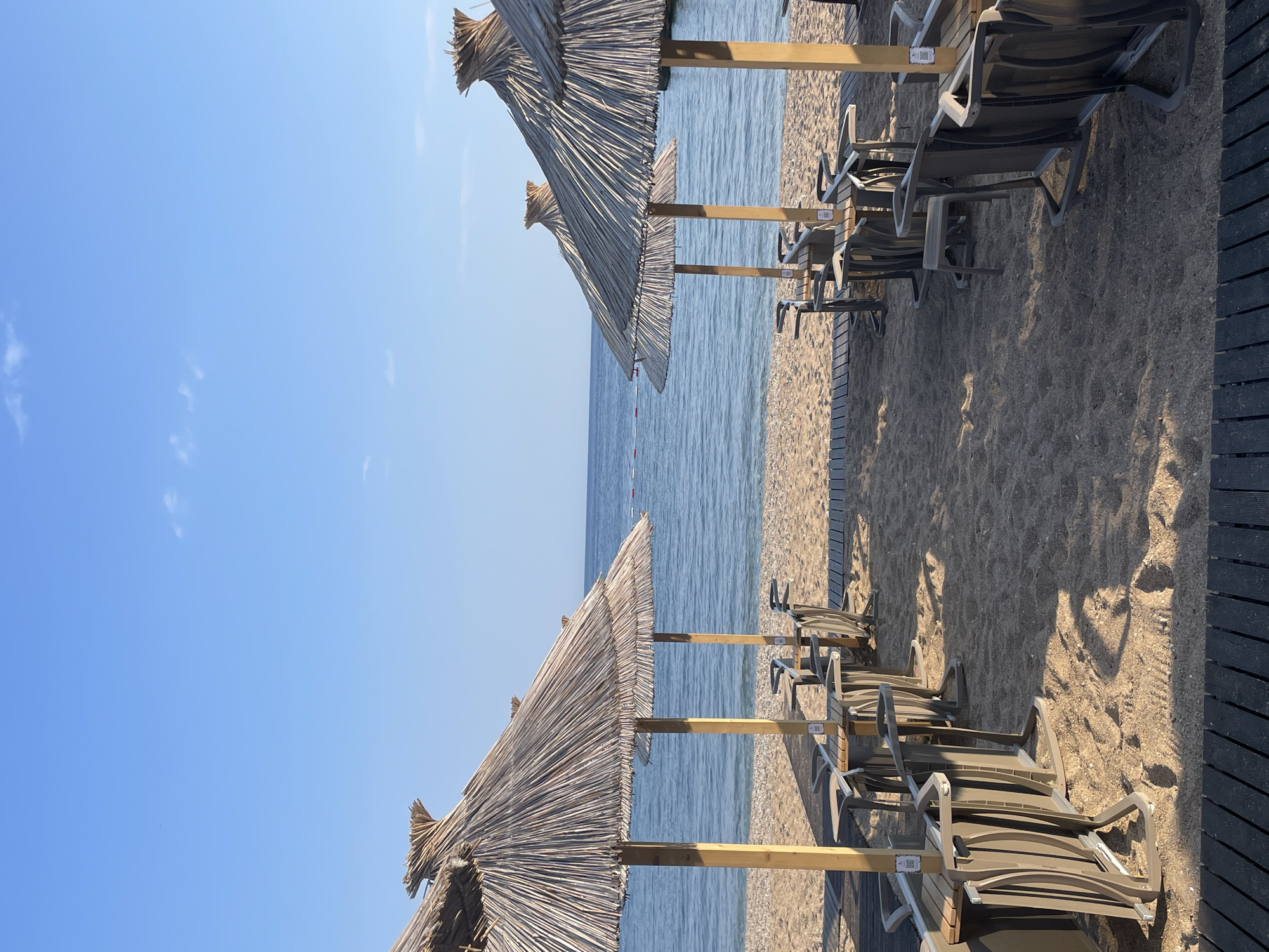 The image shows a beach scene in Budva, Montenegro. Several rows of beach chairs and straw umbrellas are lined up on the sand near the water. The sky is clear and blue. The water appears calm. A wooden walkway is visible on the right side of the image. There are no people visible in the scene.