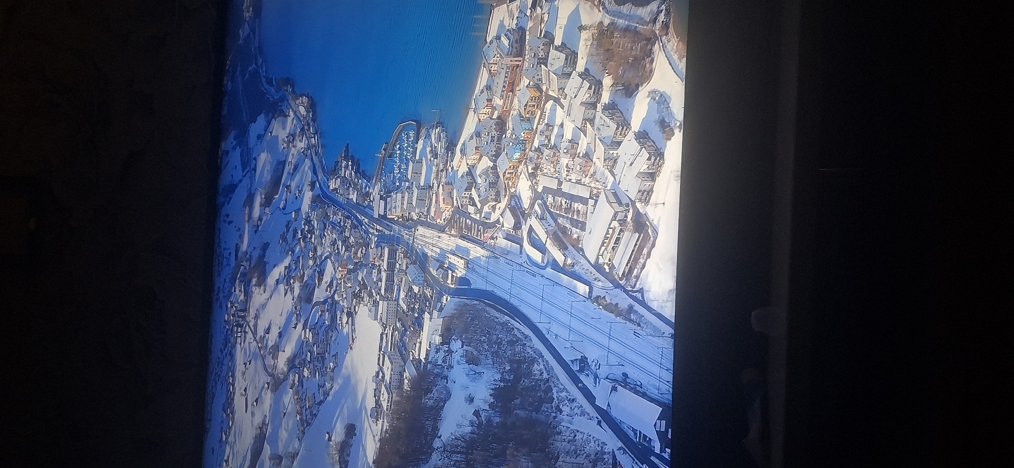 This is an aerial drone shot looking down on a snowy landscape with a body of water and a town. The town has many buildings with pitched roofs covered in snow. There is a set of train tracks running through the town. A marina with several boats is visible along the water's edge. The water is a deep blue. The ground and hills are covered in snow. The scene is brightly lit, suggesting daytime. The location is NOT Bila Tserkva, Ukraine.