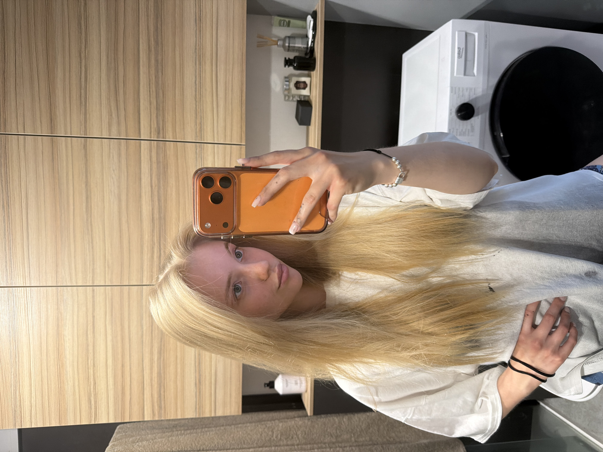 A young woman with blonde hair and light complexion is taking a selfie with a smartphone. The phone has an orange case. She's wearing a white t-shirt and has bracelets on both wrists. The background shows a light wood-grain cabinet, a shelf with various small objects, and a white washing machine or dryer with a black door. The location is likely an indoor space, possibly a bathroom or laundry room. The city and state are unknown.
