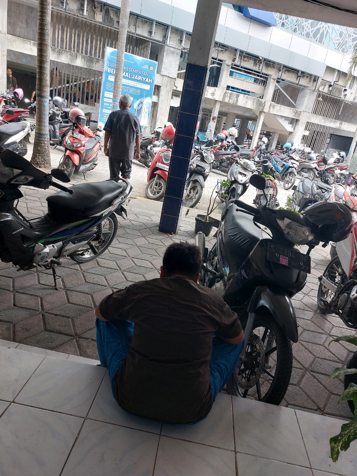 The image shows a busy motorcycle parking area in front of a building, likely an office or public facility, in Sumberrejo, Indonesia. A man in a brown shirt and blue pants is sitting on the ground, facing away from the camera, near a black motorcycle. In the background, several other motorcycles are parked, some with riders or people standing nearby. The building features concrete pillars and a metal fence. A large blue and white sign with Indonesian text is visible on the building's facade. The ground is paved with interlocking concrete blocks and white tiles. The scene appears to be during daylight hours.