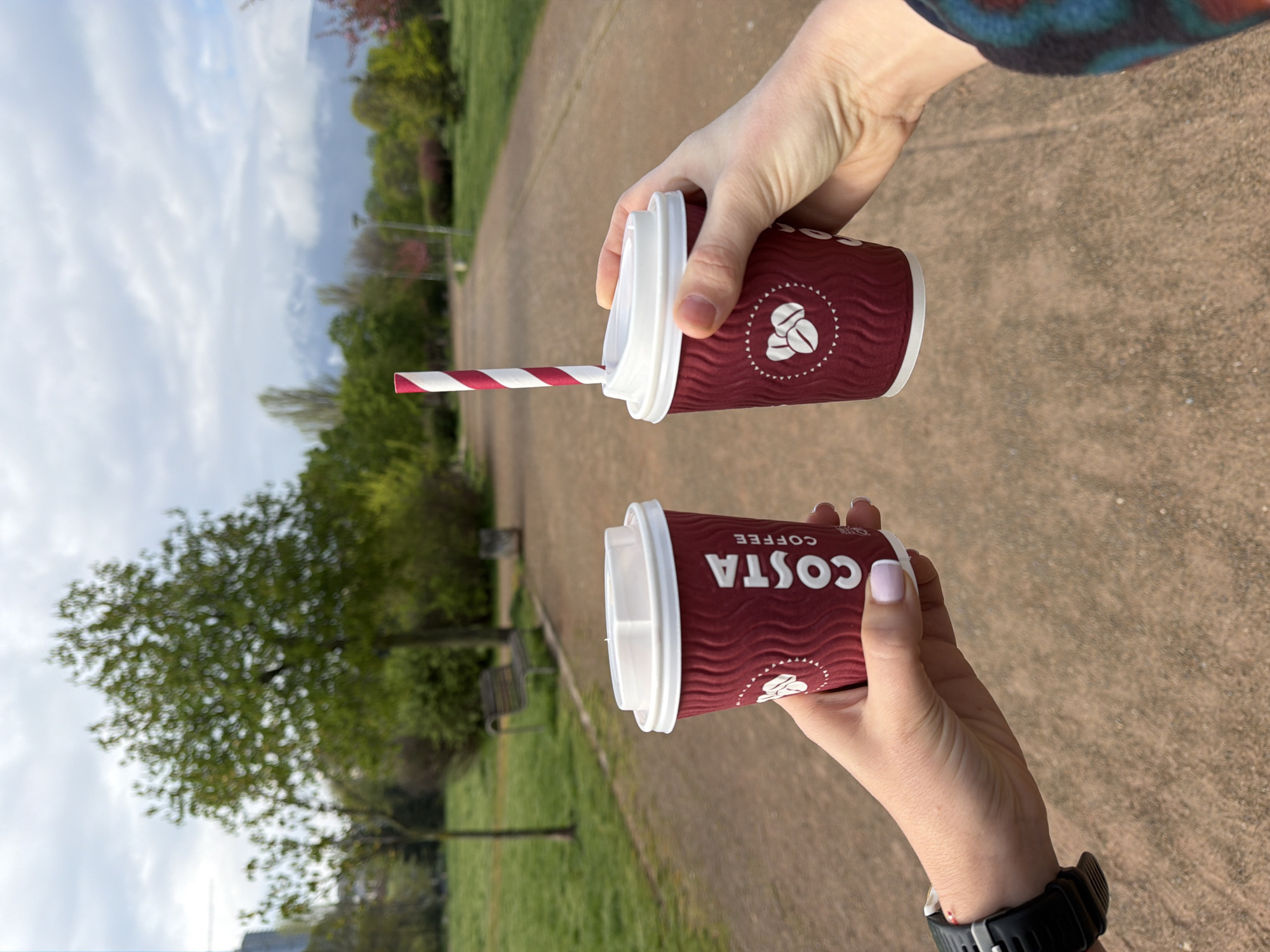 The image captures two hands holding Costa Coffee cups outdoors, likely in a park in Sofia, Bulgaria. The setting features a wide, light-colored paved path, flanked by green grass and numerous trees with vibrant green foliage. In the background, a cloudy sky is visible above the treeline. Park benches can be subtly seen along the path to the left.
The upper hand holds a dark red Costa Coffee cup with a white lid, from which a red and white striped paper straw emerges. This hand appears bare, without nail polish, and a multi-colored garment is visible near the wrist. The cup displays the Costa Coffee logo, featuring three coffee beans within a circular design, and the brand name "COSTA COFFEE" printed in white.
The lower hand holds a second identical Costa Coffee cup. This hand features light pink nail polish and wears a black wristwatch with a silver buckle on the wrist. The "COSTA COFFEE" text is clearly visible and oriented correctly on this cup.
The scene suggests two individuals are enjoying hot beverages while strolling or resting in the park. The time of day is daytime, under what appears to be an overcast or partly cloudy sky. The image conveys a casual, leisurely moment spent outdoors.