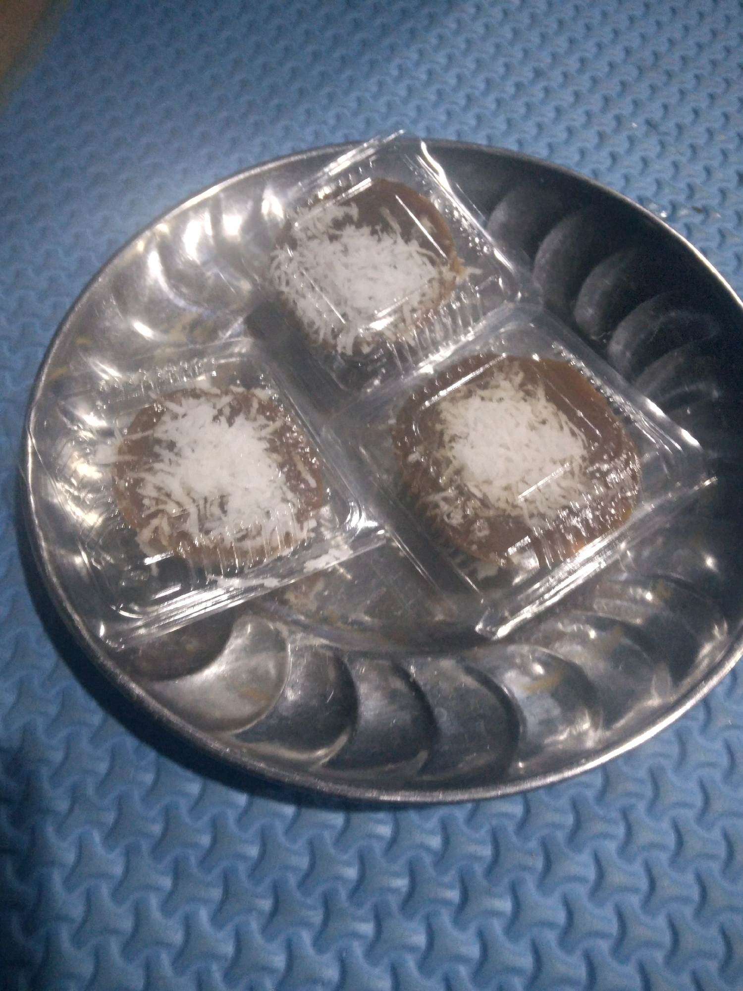 The image shows three small, individually packaged desserts topped with shredded coconut, resting on a silver-colored, ridged platter. The platter is placed on a blue, textured surface. The desserts appear to be brown in color and are in clear plastic packaging. The lighting suggests an indoor setting, possibly a home kitchen or a small eatery. The overall scene depicts a simple presentation of food. There is no visible indication of the specific location within Batam, Indonesia.