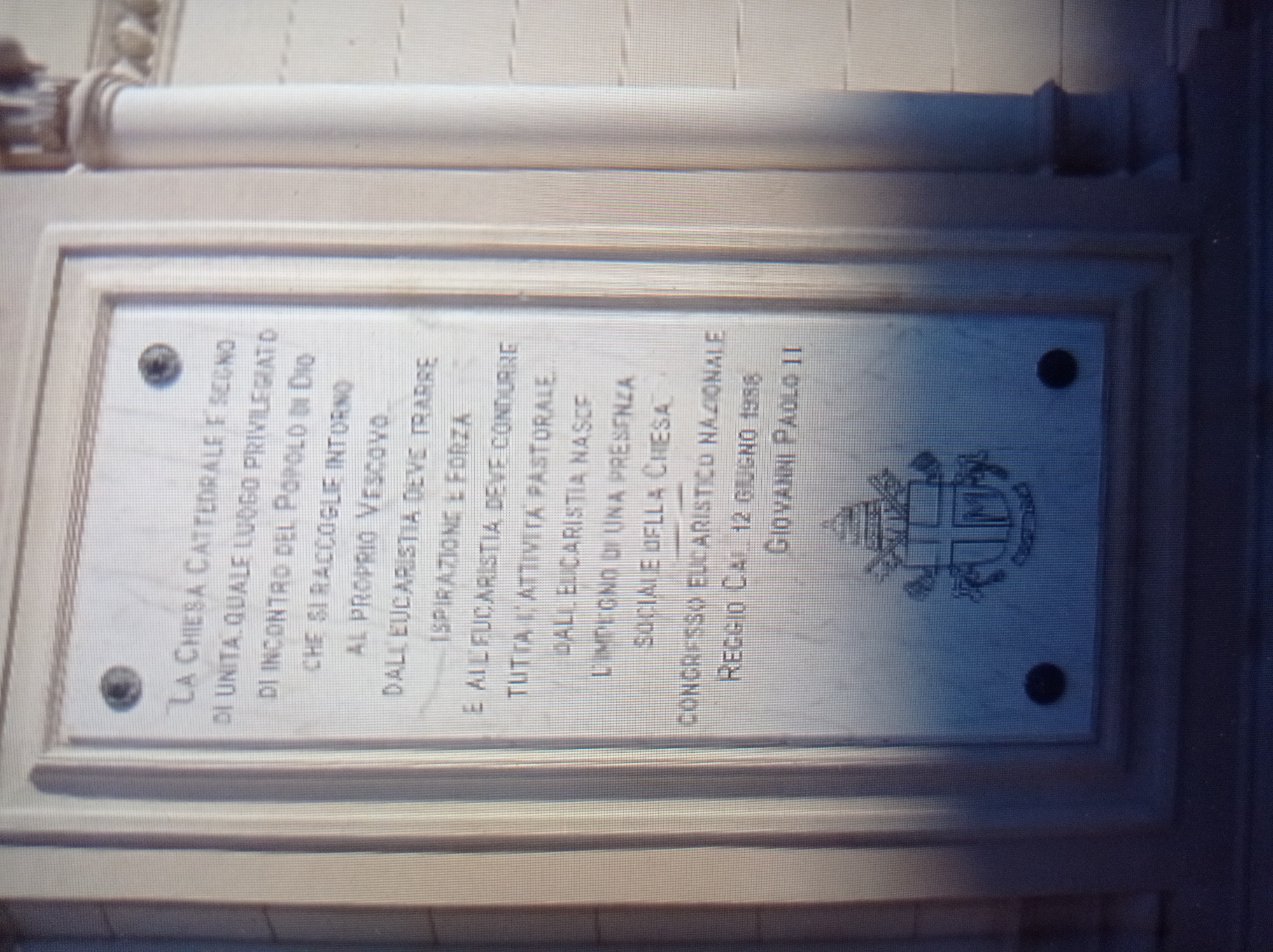 The image shows a stone plaque mounted on the exterior of a building. The plaque is rectangular with a decorative border and contains an inscription in Italian. The inscription reads: "LA CHIESA CATTEDRALE E SEGNO DI UNITA, QUALE LUOGO PRIVILEGIATO DI INCONTRO DEL POPOLO DI DIO CHE SI RACCOGLIE INTORNO AL PROPRIO VESCOVO. DALL'EUCHARISTIA DEVE TRARRE ISPIRAZIONE E FORZA E ALL'EUCHARISTIA DEVE CONDURRE TUTTA L'ATTIVITA PASTORALE. DALL'EUCHARISTIA NASCE L'IMPEGNO DI UNA PRESENZA SOCIALE DELLA CHIESA. CONGRESSO EUCARISTICO NAZIONALE REGGIO CAL. 12 GIUGNO 1958 GIOVANNI PAOLO II". Below the text, there is a coat of arms, likely that of the Pope. The plaque is situated on a wall that appears to be made of light-colored stone, with vertical lines suggesting architectural features like columns or pilasters. The lighting suggests it is daytime. There are no people or animals visible in the image. The image quality is moderate, with some pixelation and slight blur. The provided location, the city of Mukachevo, Ukraine, is not supported by any visual evidence in the image itself. The text on the plaque mentions "REGGIO CAL.", which likely refers to Reggio Calabria, Italy, and "GIOVANNI PAOLO II", Pope John Paul II, who visited Italy multiple times, including Reggio Calabria in 1988.