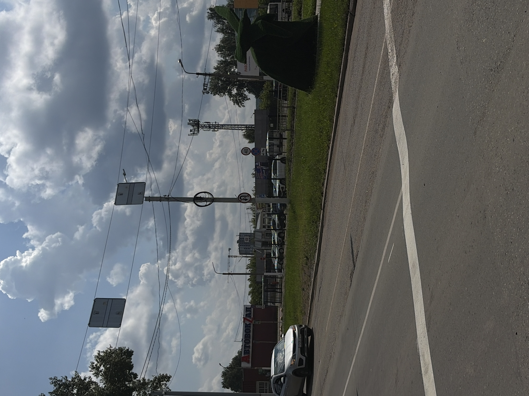 The image depicts a street scene with a clear sky and scattered clouds. On the left side of the frame, a silver car is driving on the asphalt road, which is marked with white lines. In the background, there is a building with "АВТОВОКЗАЛ" (Bus Station) written on it in large red letters. Several other buildings, a fence, and trees are also visible. To the right of the frame, there is a large, green, sculpted bush resembling a figure, possibly a rabbit or a gnome, on a grassy area. Further in the background on the right, a cell tower and a street lamp are present. The overall environment appears to be an urban setting.