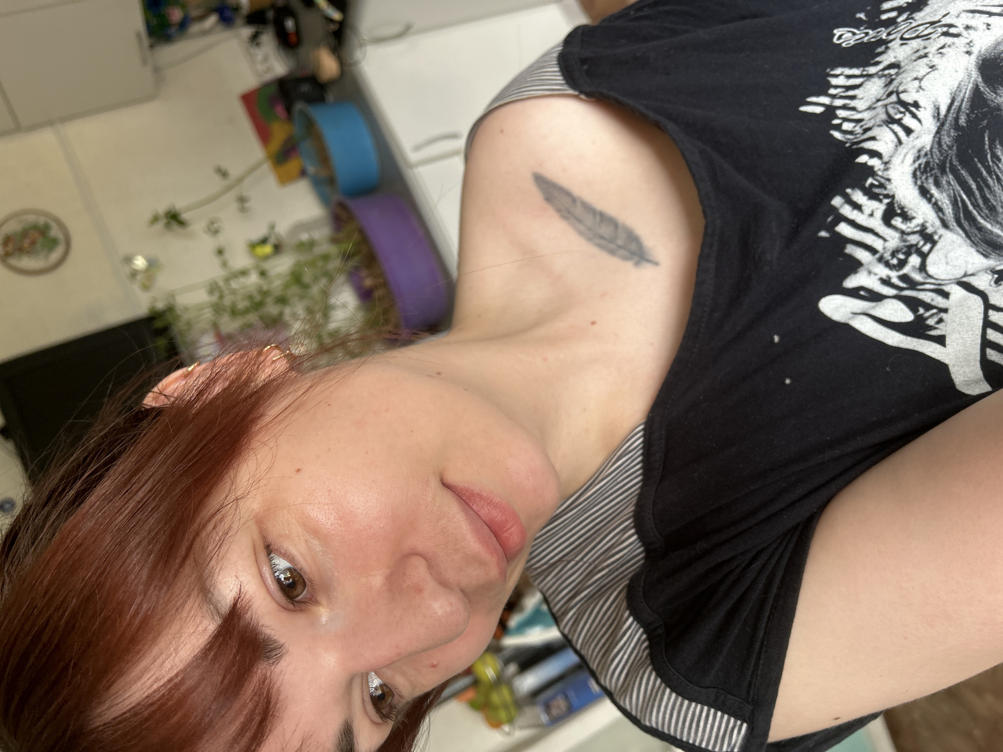 A selfie-style image, rotated, showing a woman with auburn hair. She is wearing a black t-shirt with a white graphic design. A feather tattoo is visible on her upper arm. The background includes a blurred view of a room with various objects and a glimpse of outdoor greenery. This image does not depict a location in the city of Фоки, Russia.