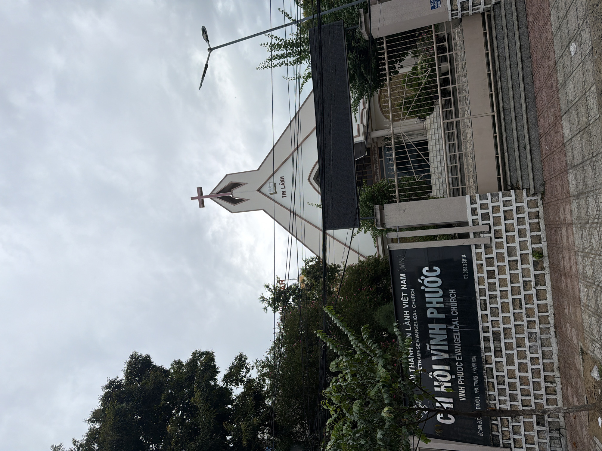 The image shows the front of a Vietnamese Evangelical Church in Nha Trang, Vietnam. The church building is white with a prominent cross at the top of its pointed roof. The name of the church is displayed on a black sign in both Vietnamese and English: "CHI HỘI VĨNH PHƯỚC VINH PHUOC EVANGELICAL CHURCH". Below this, there is text indicating the address: "DC: 694 ĐUC. THÁNG 4 - NHA TRANG - KHÁNH HÒA". The church is surrounded by trees and a metal fence with a gate. The sky is overcast, and power lines are visible in the foreground. There are no people or animals visible in the image.