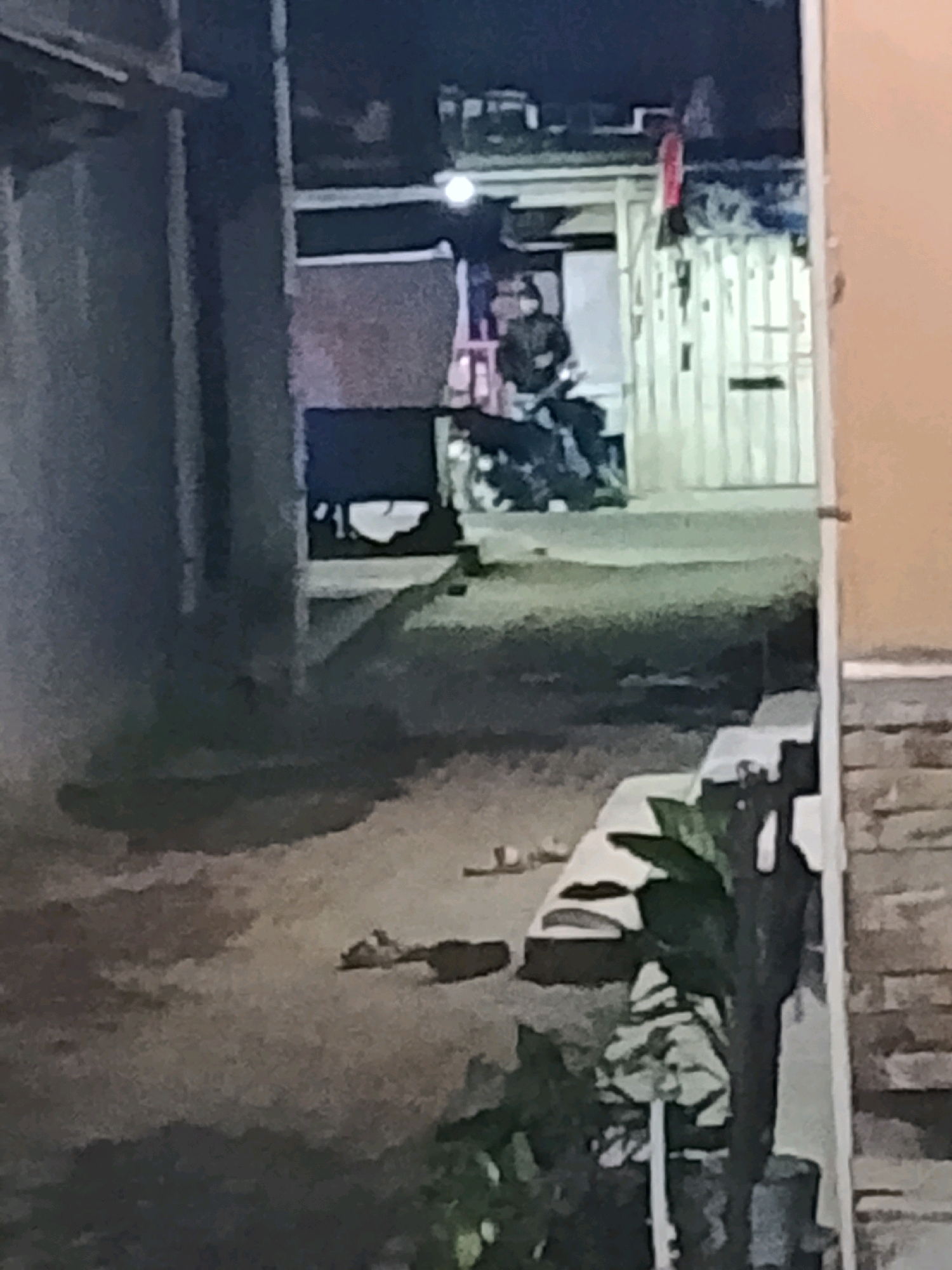 A nighttime, low-light, slightly blurry, medium shot captures a narrow alleyway or side street in the city of Ujungrusi, Indonesia. The ground appears to be paved with rough concrete or asphalt. In the foreground, several pairs of sandals are scattered on the ground. To the right, a portion of a building with a textured, brick-like facade is visible. A potted plant with dark green leaves is situated on a ledge in the mid-ground. In the background, a person wearing a dark jacket and a helmet is seated on a motorcycle. The motorcycle is positioned in front of a building with corrugated metal siding. The overall impression is one of a dimly lit, possibly residential or commercial backstreet.