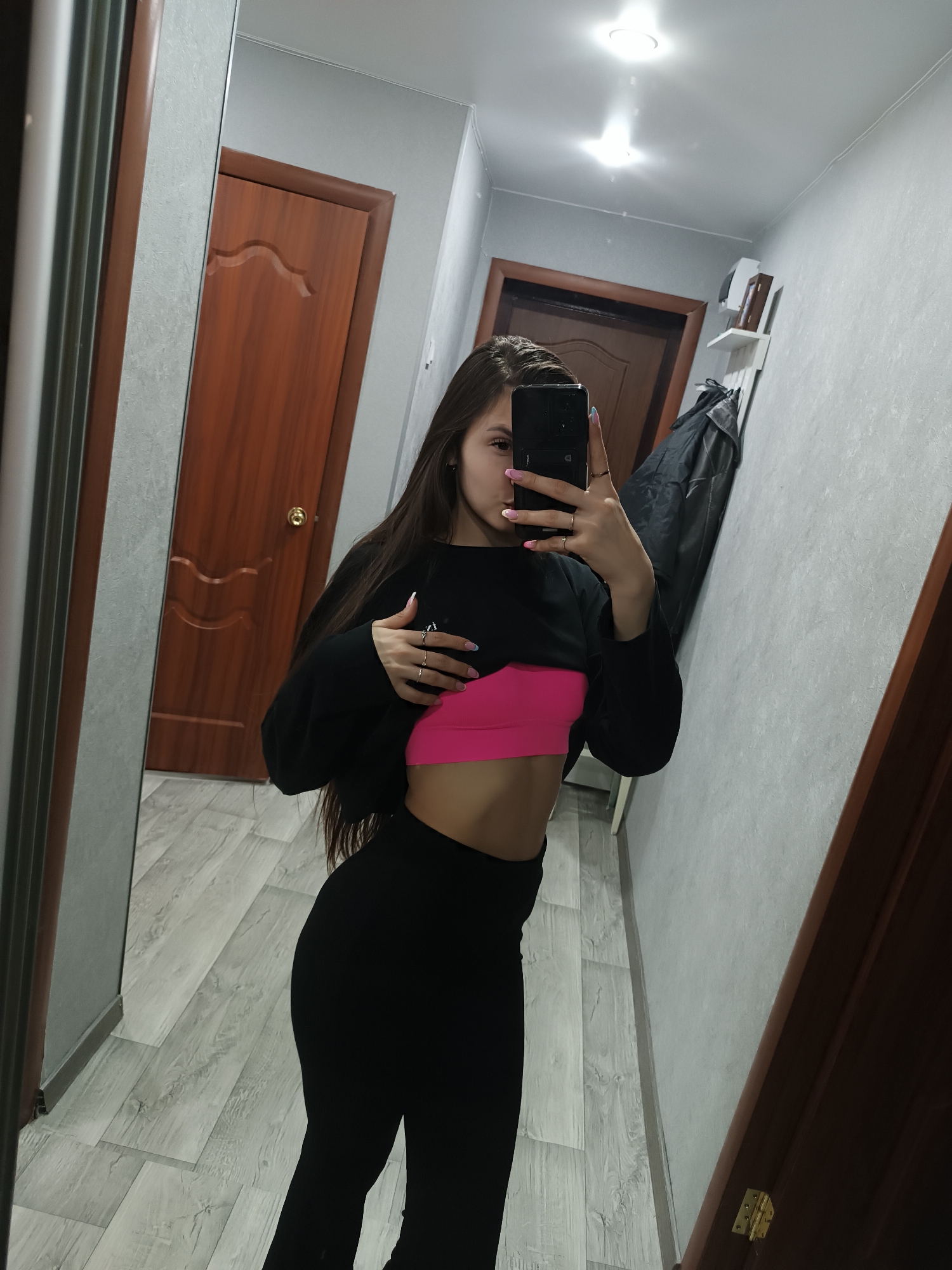 A woman is taking a selfie in what appears to be an apartment hallway. She is wearing a black long-sleeved shirt, a pink sports bra, and black leggings. The hallway has light gray wood-patterned flooring, a brown door, and light gray walls. A mirror is visible on the left side of the image, and a coat rack with a black garment is seen at the end of the hallway. The location is Chaikovsky, Russia.