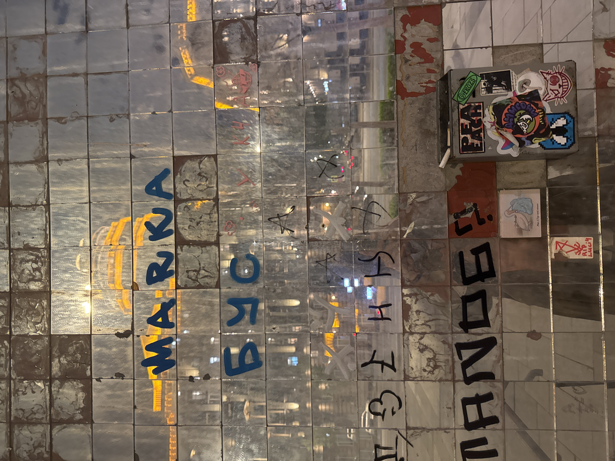 The image shows a wall covered in small, reflective tiles in what appears to be Moscow, Russia. The tiles are adorned with graffiti, stickers, and handwritten messages. The word "MARKA" is written vertically in blue paint. Other messages and symbols are also visible. The tiles reflect the city's surroundings, including buildings and streetlights. A box with various stickers is affixed to the wall. The wall looks aged and weathered. The image is taken at night or in low-light conditions.