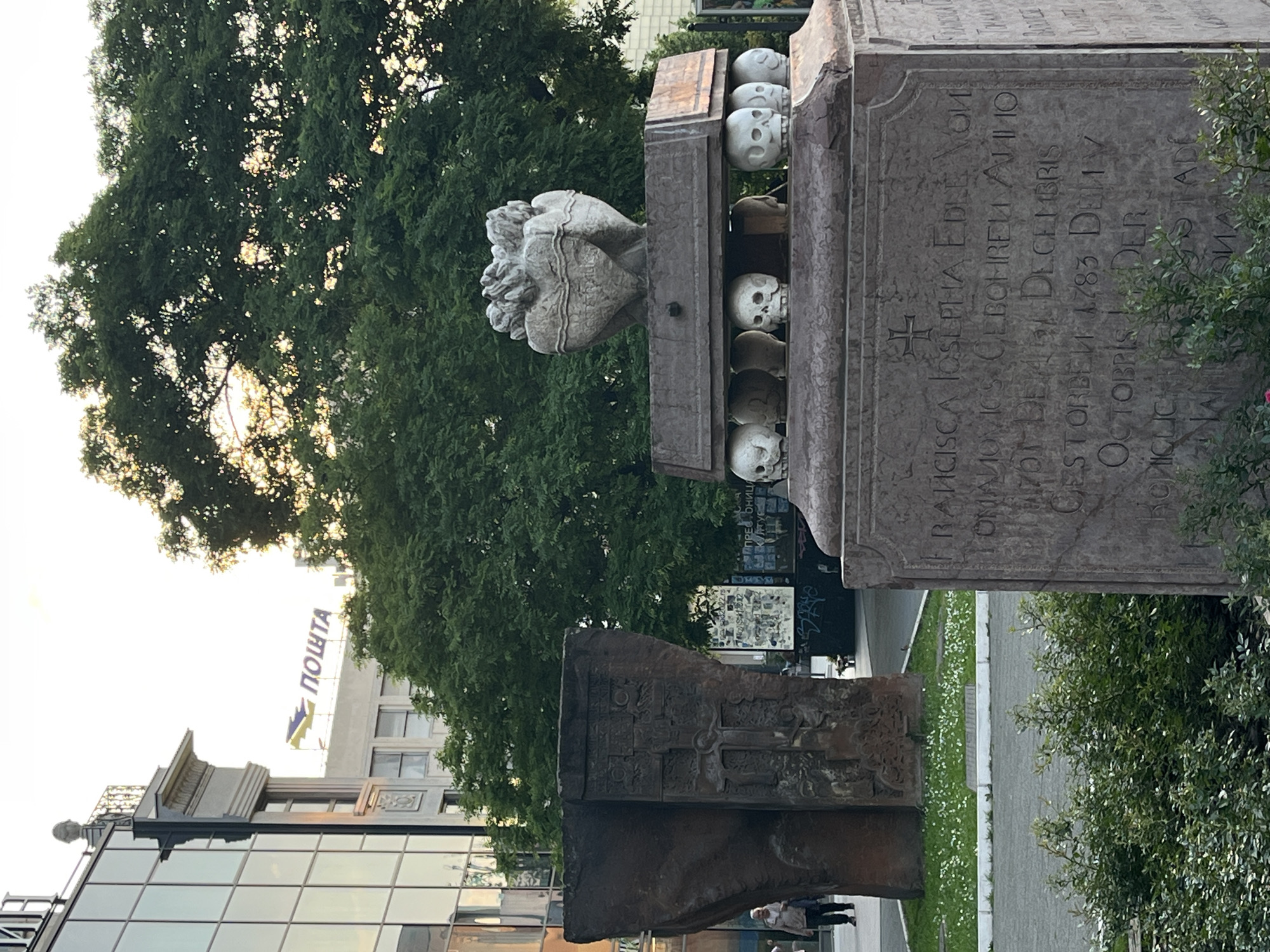 This image depicts the Plague Column in Novi Sad, Serbia, a monument commemorating the victims of the plague epidemics of the 18th century. The column, made of stone, features skulls on its upper sections. An inscription is visible on one of the sides. In the background, a building with the Cyrillic word "ПОШТА" (Post office) is seen, indicating the city location. A tree stands behind the column and a modern building with glass windows is reflected in the foreground.