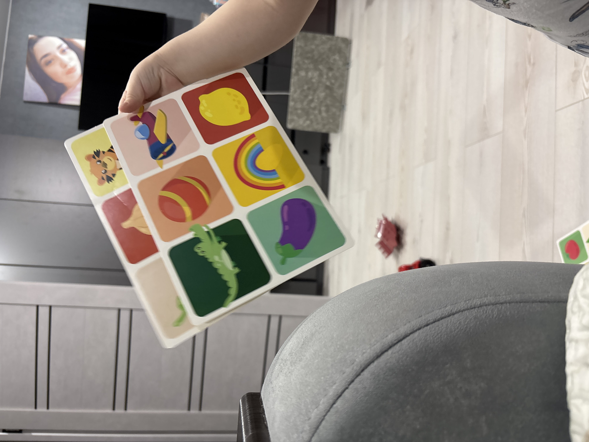 A close-up shot shows a child's hand holding a set of educational picture cards. The cards display images of various fruits, vegetables, and objects such as a lemon, eggplant, rainbow, ball, and tiger. The cards are held in front of a blurred background that includes a grey chair and a wall with a picture of a woman. The scene appears to be indoors, possibly in a home in the city of Chaikovsky, Russia.