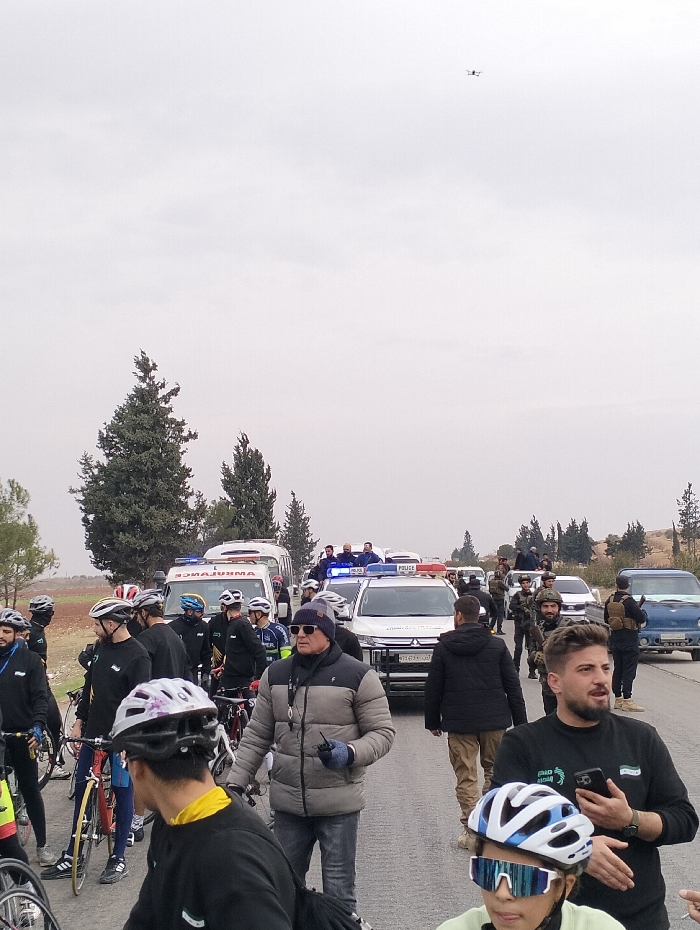 A group of cyclists in black jerseys with helmets are gathered on a road with several support vehicles including police cars and an ambulance. Armed security personnel are visible. One man stands out in a grey puffer jacket. A drone flies overhead. The scene conveys a sense of an organized cycling event with safety and security measures in place. The weather appears overcast.