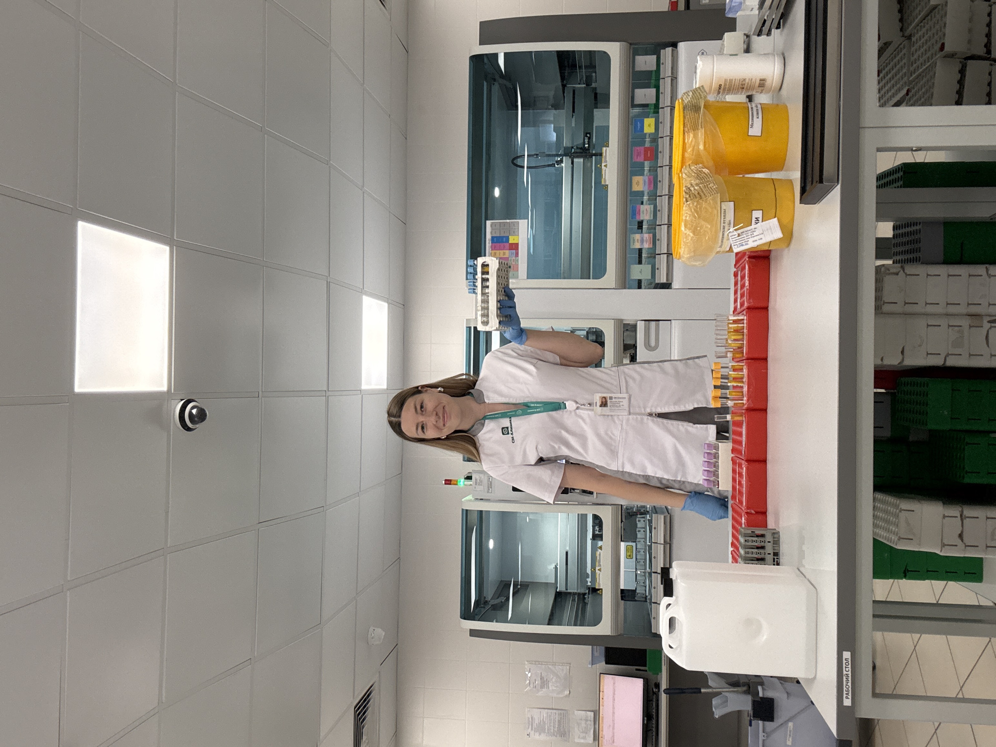 A woman in a white lab coat stands in a laboratory, holding a rack of test tubes. She is smiling and looking at the camera. Behind her are various pieces of laboratory equipment, including automated analyzers and racks of samples. The lab appears to be clean and well-organized, with fluorescent lighting and tiled walls. The woman is wearing blue gloves and has a lanyard with an ID badge around her neck. The overall impression is one of a professional and efficient scientific setting.