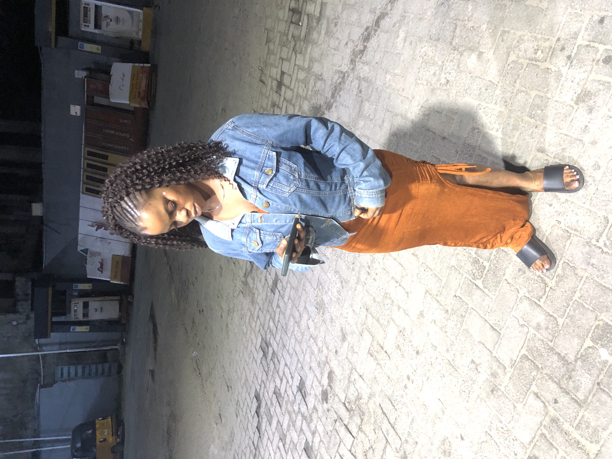 A young Black woman is standing on a brick-paved surface at night. She is wearing a denim jacket over an orange dress with a high slit and ruching on the side. She has braided hair and is looking down at her phone, which she is holding in her right hand. She is wearing black slides on her feet. In the background, there are gas pumps, a yellow three-wheeled vehicle, and various signs. The location appears to be a gas station in Ado, Nigeria. The lighting is artificial and casts shadows on the ground.