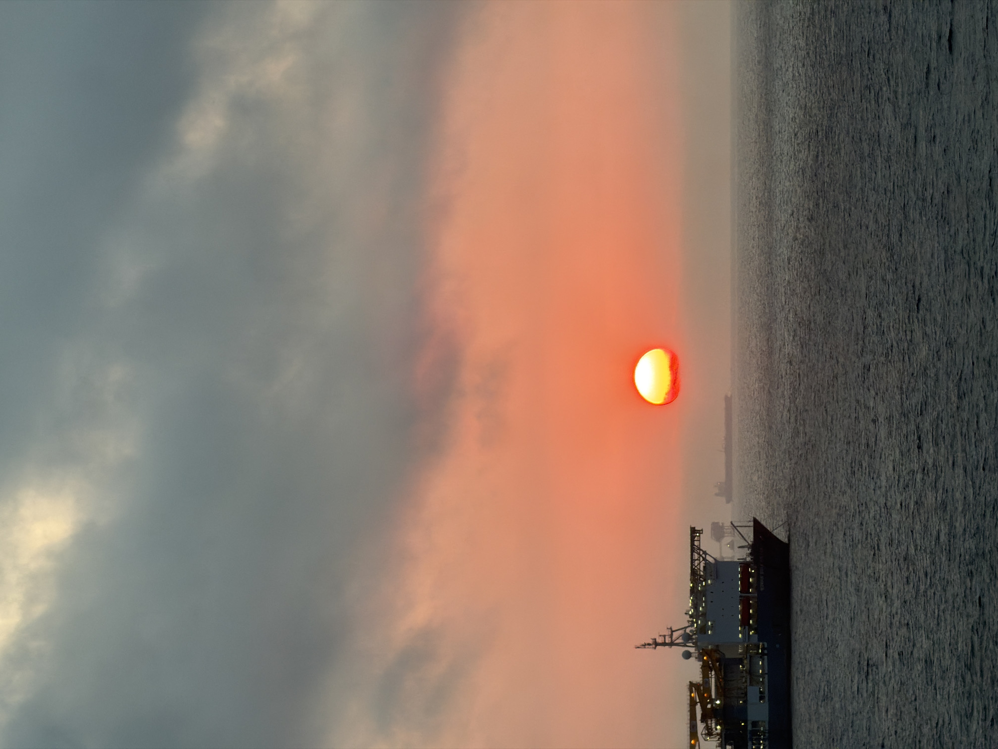 A dramatic image captures a sunset or sunrise over the ocean. The sun, a vibrant orange-red orb, is prominent on the right side of the frame, partially obscured by thick, atmospheric clouds. These clouds display a gradient of colors, from dark, cool grays and blues on the left to warm, soft pinks and oranges closer to the sun. The sky's expanse dominates the upper and left portions of the image.
The sea, dark and textured with ripples, occupies the foreground and lower right. A large vessel, possibly a cargo ship or offshore support vessel, is visible in the lower right. It features a complex superstructure, masts, and antennas, with several lights illuminated on its deck and interior, suggesting either dawn or dusk. In the far distance, another smaller or more distant ship is faintly silhouetted against the hazy horizon.
The overall scene is atmospheric and tranquil, with the low sun casting a warm glow through the cloudy sky. No specific human activity is discernible, and no text is clearly legible on the ships.
