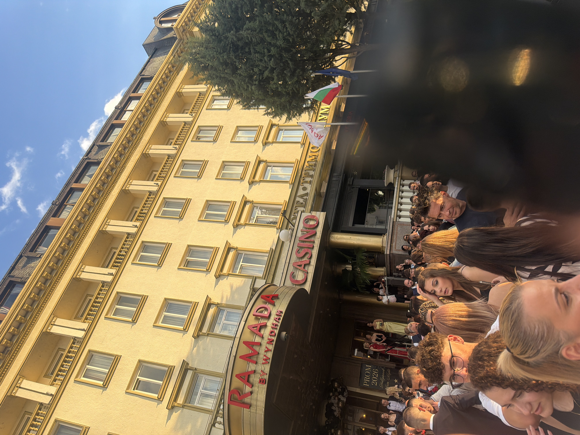 A large group of people is gathered in front of the Ramada by Wyndham Plovdiv Trimontium Hotel, in Plovdiv, Bulgaria. The hotel building is a tall, beige structure with multiple windows and balconies. The entrance is marked by "RAMADA by WYNDHAM CASINO" signage. Flags are visible above the entrance. People are standing in the hotel entrance. A sign reads "PROM 2026". The sky is blue.
