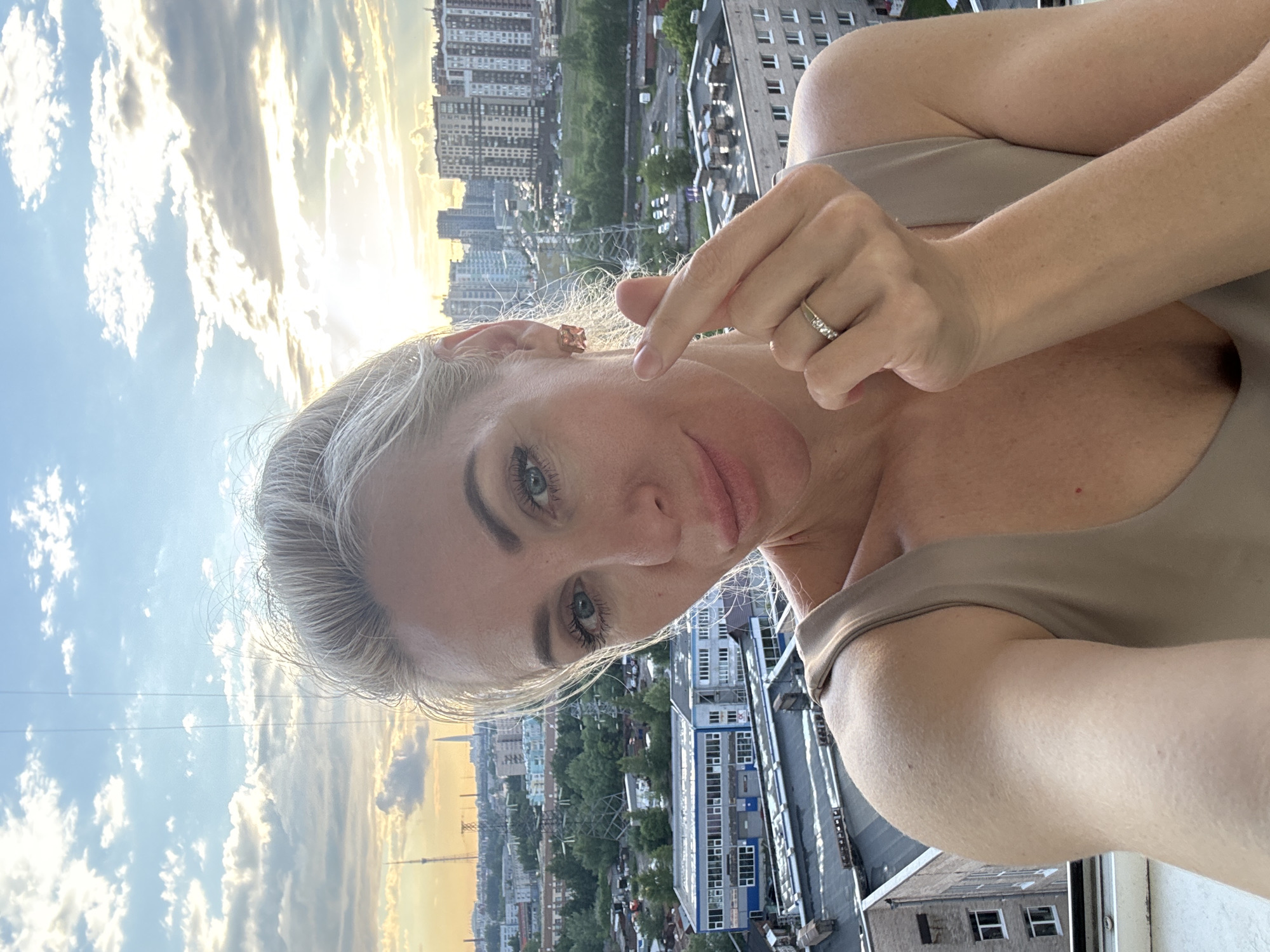 In an outdoor selfie taken on a balcony, a Caucasian woman with blonde hair pulled back and blue eyes looks towards the camera, holding her thumb and index finger together in a heart shape. She wears a beige sleeveless top and a ring with three small stones on her left hand. Behind her, a city skyline is visible under a cloudy sky with the setting sun casting a warm glow. The city appears to be St. Petersburg, Russia, indicated by the presence of the Lakhta Center skyscraper in the distance. The buildings are a mix of modern high-rises and older industrial structures, with trees and vegetation interspersed throughout. The overall scene captures a moment of casual posing against a sprawling urban backdrop at dusk.