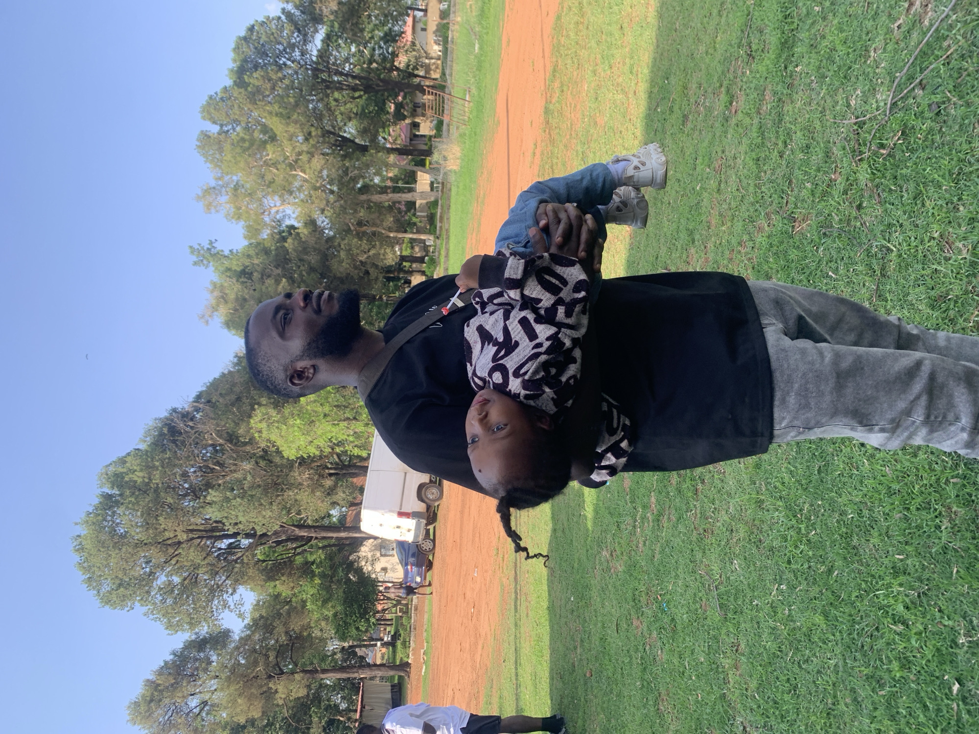 A Black man with a beard, wearing a black t-shirt and gray jeans, stands in a grassy area and holds a young Black child in his arms. The child, who is lying on their back across the man's arms, is wearing a black and white patterned sweater and blue jeans. The man is looking to his right, with the child looking directly at the camera. In the background, there is a dirt path, some trees, a white van, and a blue car. The image appears to have been taken in Jos, Nigeria, as the style of buildings, trees, and landscape are consistent with the region.