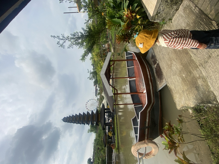 A child wearing a hat and striped shirt stands on a concrete surface near the edge of the water. A boat sits in the water, adjacent to the concrete. In the background, a tall, multi-tiered structure and a Ferris wheel are visible. Green foliage and a cloudy sky complete the scene.