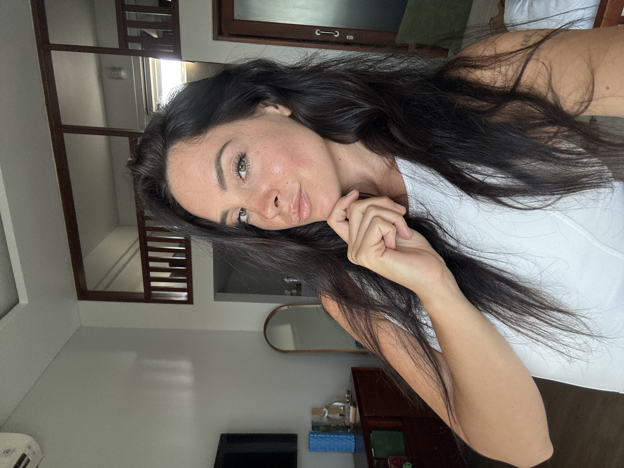 A woman with long, dark hair, wearing a white top, poses for a selfie in a room in North Kuta, Indonesia. She is looking at the camera with a slight pout, her right hand resting on her chin. The room appears to be a bedroom with a television on the left, a dresser with decorative items, and a large mirror. In the background, there is a mezzanine level with wooden railings and a door. The lighting suggests it is daytime.