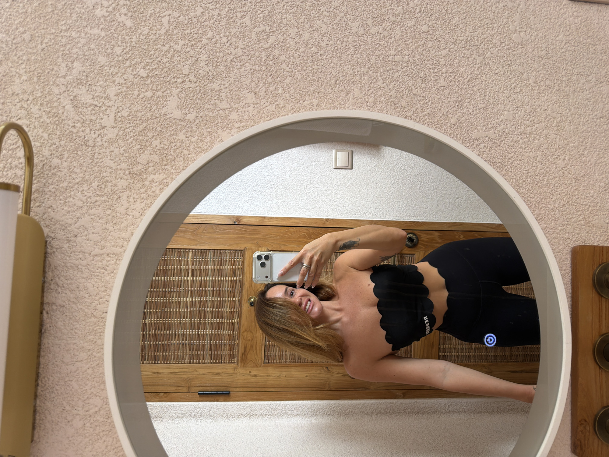 The scene is a mirror selfie taken by a woman. The woman is holding a smartphone and facing the mirror. She is wearing black leggings and a black strapless top. The mirror is round and is attached to a wall with textured, light-beige plaster. The reflection in the mirror shows a wooden cabinet with woven panels behind the woman. The setting could be a bathroom in the city of Mengwi, Indonesia.