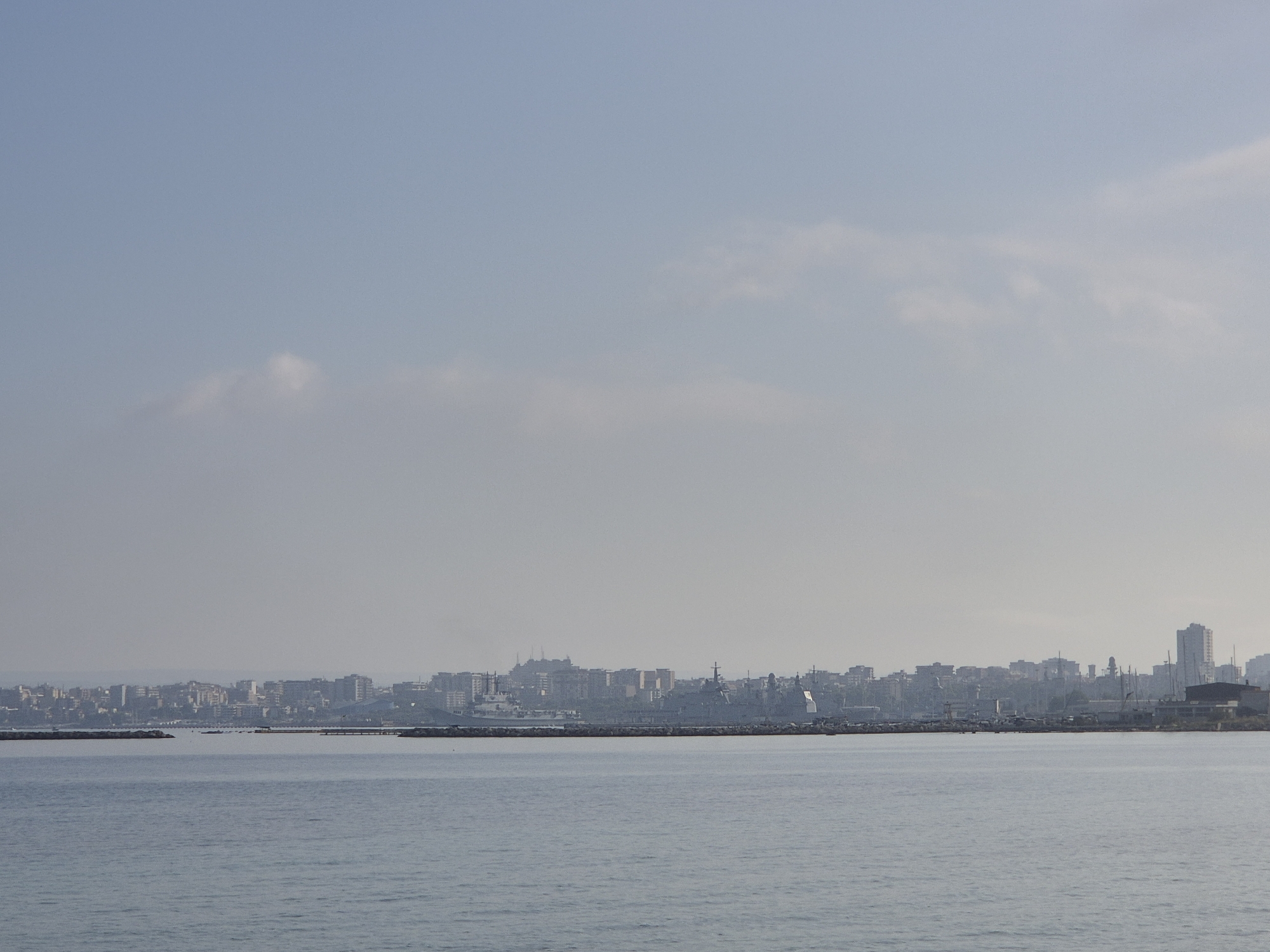 The image depicts the waterfront of Taranto, Italy, with the Mar Piccolo in the foreground and the city skyline in the background. The water is calm and a light blue, reflecting the hazy sky. In the middle ground, a breakwater, constructed of large rocks, forms a barrier between the water and the port area. Beyond the breakwater, several ships, including what appear to be naval vessels, are docked. The city skyline is a dense collection of buildings, ranging from low-rise structures to taller modern towers, with some industrial elements visible. The overall atmosphere is slightly overcast and hazy, suggesting a mild or humid day. No people are visible in the image.