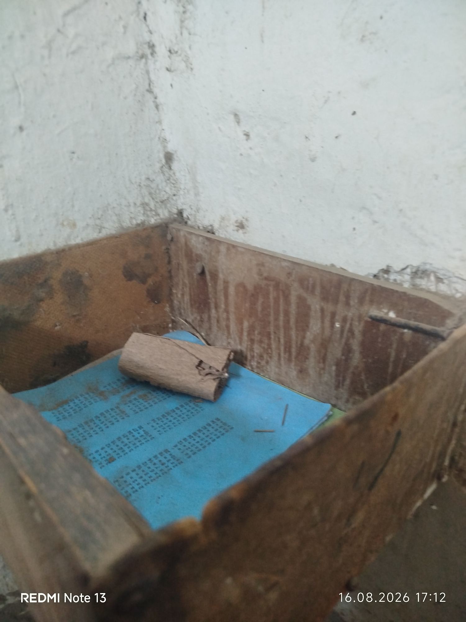 The image shows a close-up view of a dilapidated wooden box, possibly a shelf or drawer, in a dusty, unfinished room. The interior of the box is lined with a blue material that appears to be paper or fabric, upon which lies a rolled-up piece of brown, textured material, possibly a scroll or a piece of bark. The wooden structure itself is weathered and shows signs of age, with visible nails and rough surfaces. The surrounding walls are a pale, textured surface, with some dirt and discoloration. The lower left corner of the image displays the text "REDMI Note 13", and the lower right corner shows the date and time "16.08.2026 17:12". The scene is consistent with an indoor, possibly abandoned or storage area, in a building.