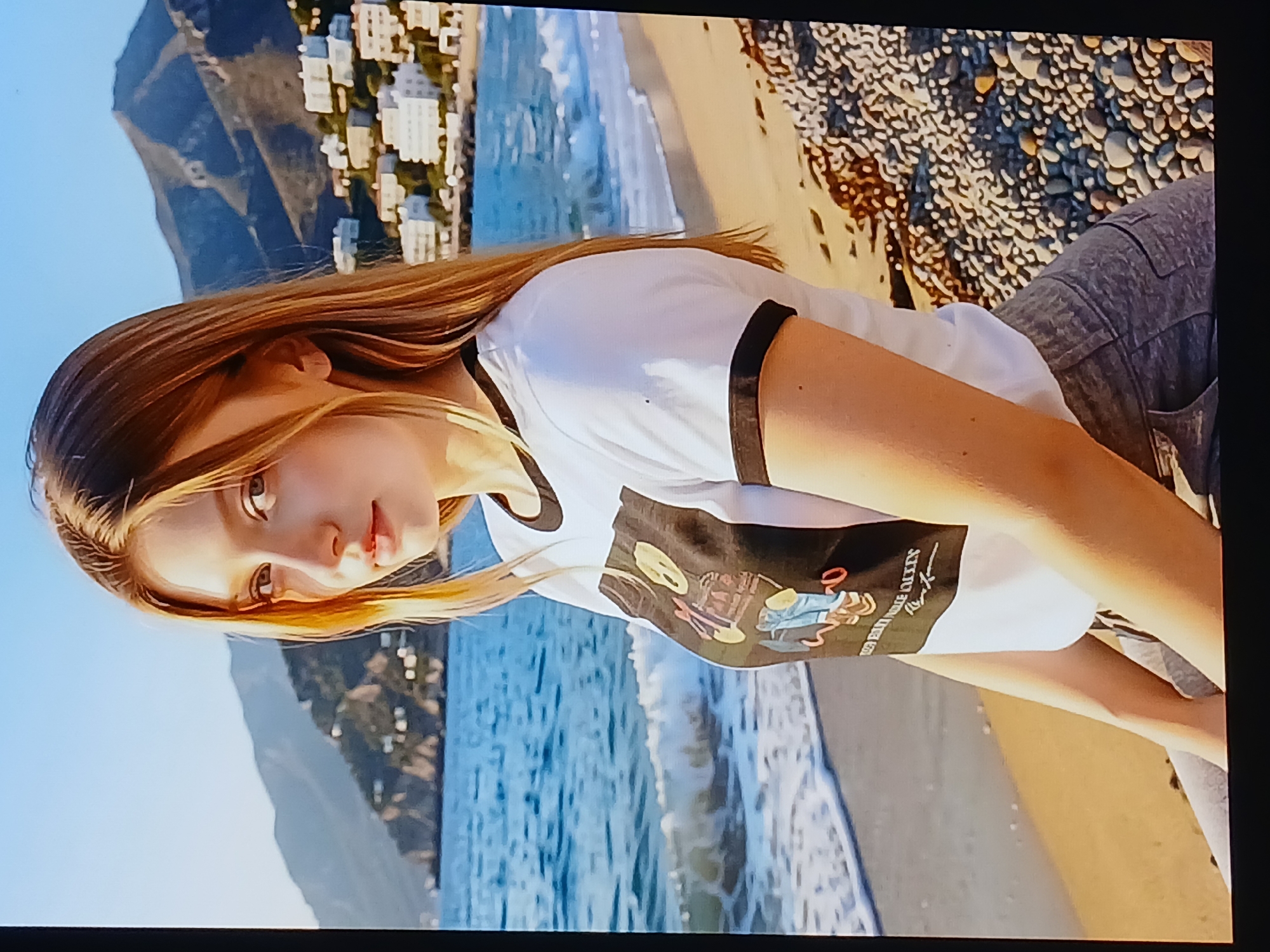 The image shows a young woman with long, blonde hair sitting on a beach with the ocean and mountains in the background. She is wearing a white t-shirt with a graphic print and grey pants. The beach is covered in pebbles and sand. The ocean is blue with waves. In the background, there are mountains with buildings on their slopes. The sky is clear and blue. The scene appears to be a coastal landscape. There is no identifiable information in the image that suggests the location is the city of Dnipro, Ukraine.