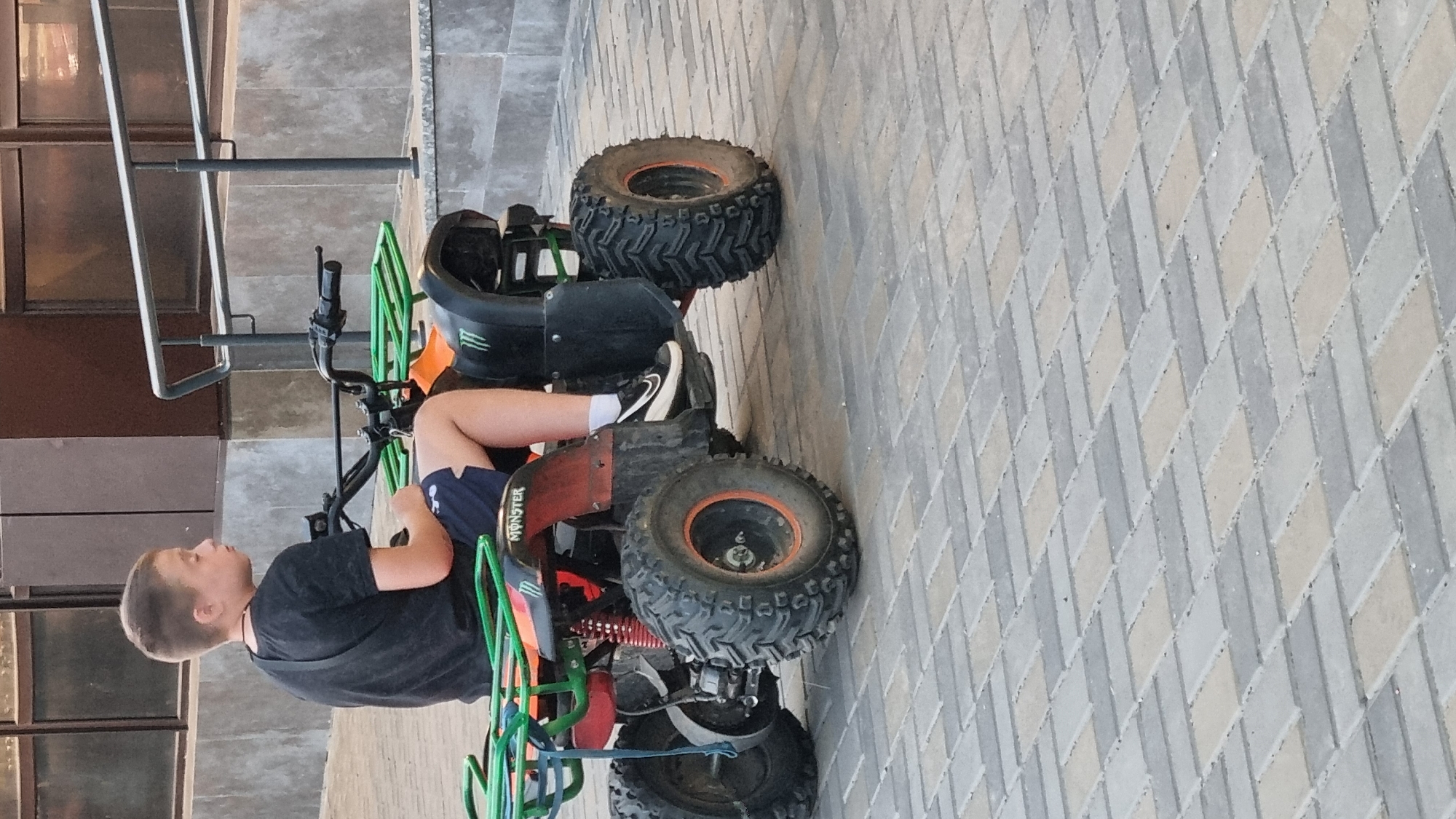 In Smila, Ukraine, a young boy in a black t-shirt and dark shorts rides a red and black ATV with green racks. The ATV has "MONSTER" branding on its side. The boy is wearing white socks and black athletic shoes. He is positioned on the ATV, holding the handlebars. The scene takes place outdoors on a paved area with a brick-like pattern. In the background, a building with dark windows and a metal railing is visible. The overall lighting suggests it is daytime.
