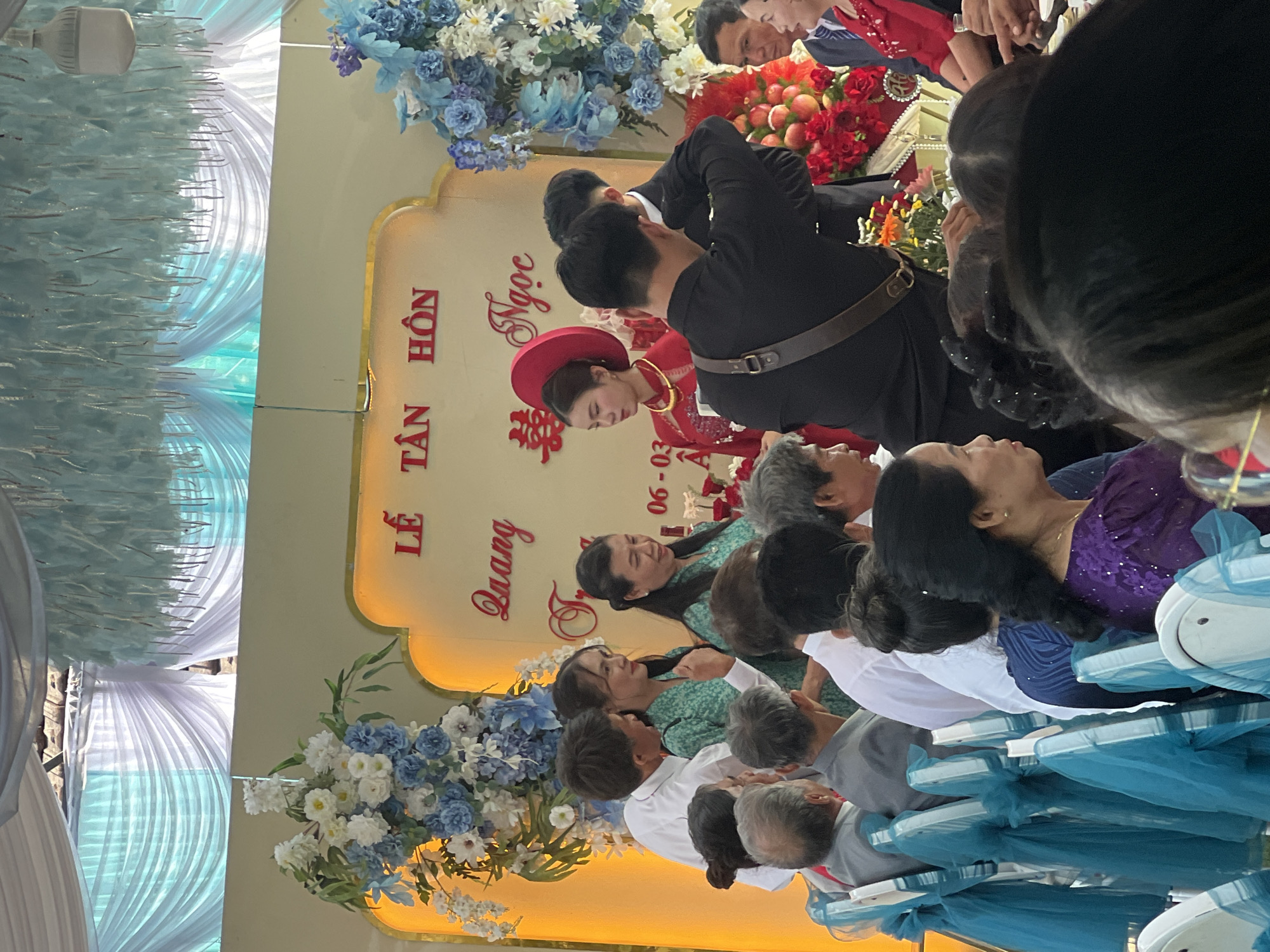 This image captures a vibrant wedding or engagement ceremony in Bình An, Vietnam. The scene is set in a decorated event space, possibly indoors or under a tent, adorned with white and light blue draping that creates an elegant backdrop.
The focal point is a woman, likely the bride, dressed in a traditional red Vietnamese áo dài and khăn đóng (conical hat), standing near a decorative golden arch. She is seen from behind, interacting with others. Standing close to her is a man in a black suit, also facing away from the camera. Surrounding them are numerous guests, some seated on chairs decorated with light blue sashes, and others standing, observing the ceremony. Among the guests, two women in green dresses are clearly visible in the mid-ground.
The golden arch, serving as a ceremonial backdrop, is illuminated and features prominent Vietnamese text. "LỄ TÂN HÔN" translates to "Wedding Ceremony." Below this, the names "Quang" and "Ngọc" are displayed, likely referring to the bride and groom. A double happiness symbol (囍) is also present, common in weddings. A date, "06 - 03," is visible, possibly indicating March 6th or June 3rd. The arch is flanked by elaborate floral arrangements featuring white, blue, and some red flowers, along with arrangements of red fruits.
The atmosphere appears festive and celebratory, with guests gathered for the special occasion. The lighting suggests it is daytime or a brightly lit indoor venue.