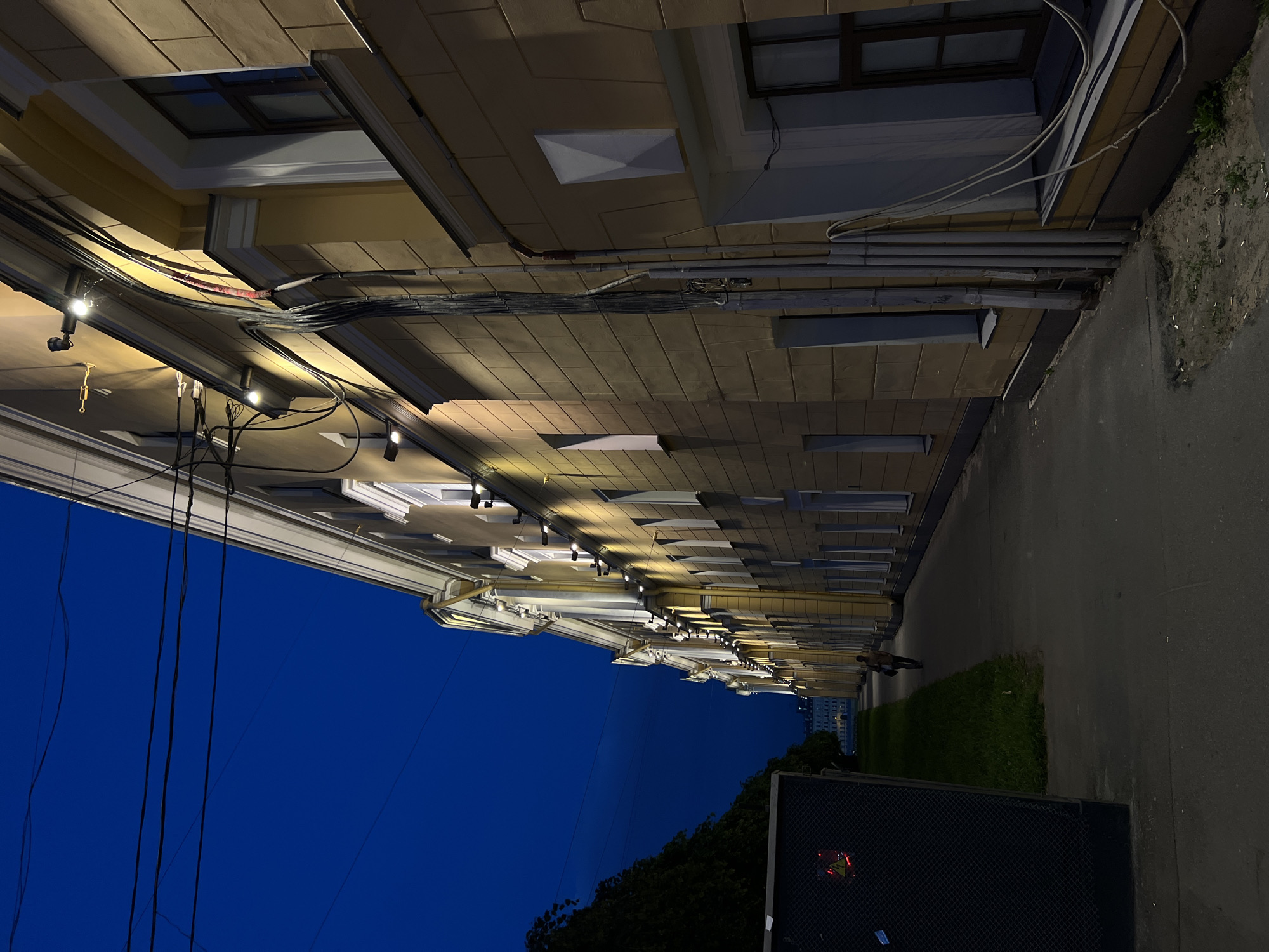 The image depicts a dimly lit street scene at dusk or night in Saint Petersburg, Russia, likely near a historical building. On the right side of the frame, a long, yellow building stretches into the distance, its facade illuminated by strings of warm-toned lights mounted along the cornice. The building has multiple windows, some dark and some showing interior light. Exposed wiring and conduits run vertically along the facade.
To the left of the building, a paved walkway runs parallel to it, leading towards the vanishing point. A lone pedestrian, seen from behind and walking away from the camera, is positioned in the middle distance on the walkway. To the far left, a dark, netted enclosure or structure is visible, featuring a red warning sign with a lightning bolt symbol. Beyond this enclosure, dark trees line the edge of the scene. The sky is a deep, clear blue, characteristic of twilight. The overall atmosphere is calm and somewhat solitary.