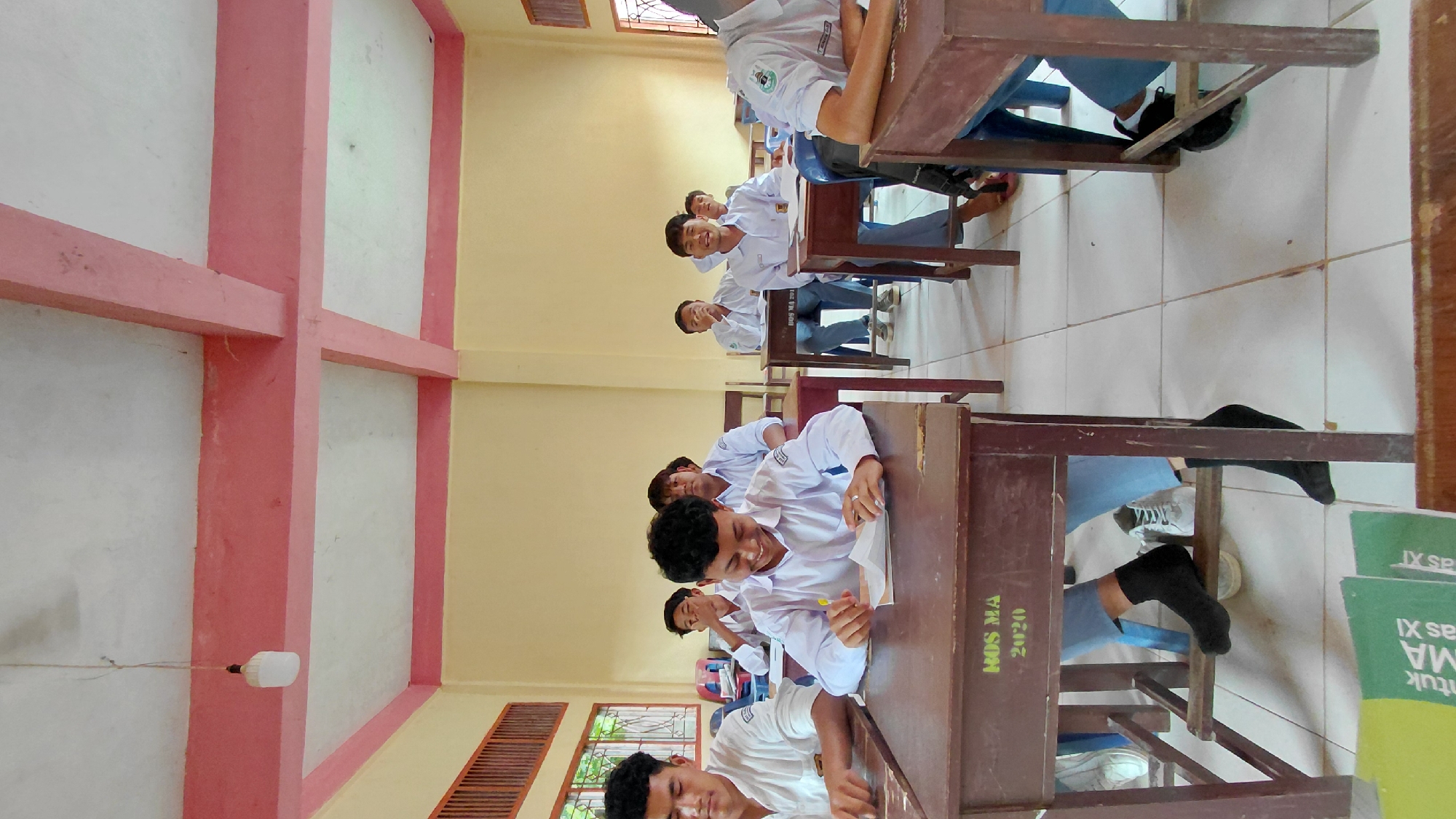 The image shows a classroom in Lhokseumawe, Indonesia. Several students, dressed in white school uniforms, are seated at individual wooden desks arranged in rows. Some students are writing or looking at papers on their desks, while others are looking towards the camera. The classroom has tiled floors, walls painted in pale yellow and white with pink trim, and windows. A light fixture hangs from the ceiling. Some of the desks have textbooks or notebooks underneath, and the desks are labeled 'MOS MA 2020'.