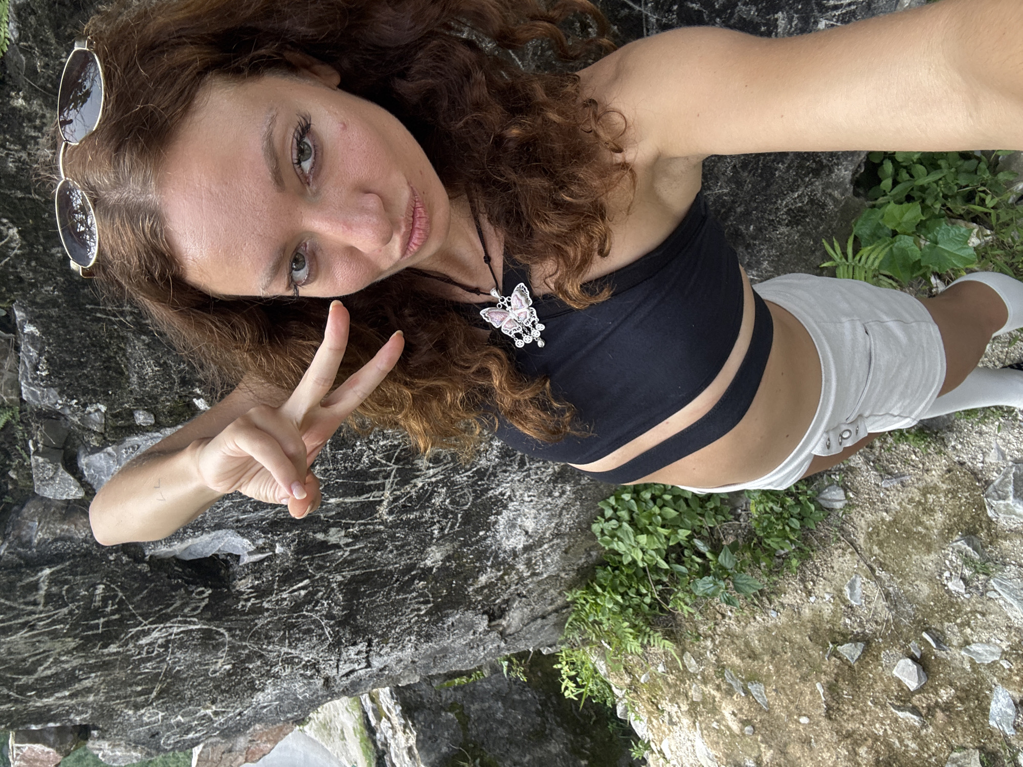 A woman is seen in a selfie in Đường Thượng Commune, Vietnam, looking directly at the camera. She holds up a peace sign with her left hand. She has long, curly brown hair and is wearing sunglasses on her head. She is wearing a black strapless top and white shorts. A necklace with a butterfly pendant is around her neck. She is standing on a textured gray rocky surface. Green plants are visible in the lower right corner of the image. The ground in the lower right corner has a light brown, dirt-like quality, with patches of green vegetation and small stones. There is graffiti etched into the rock to the left.