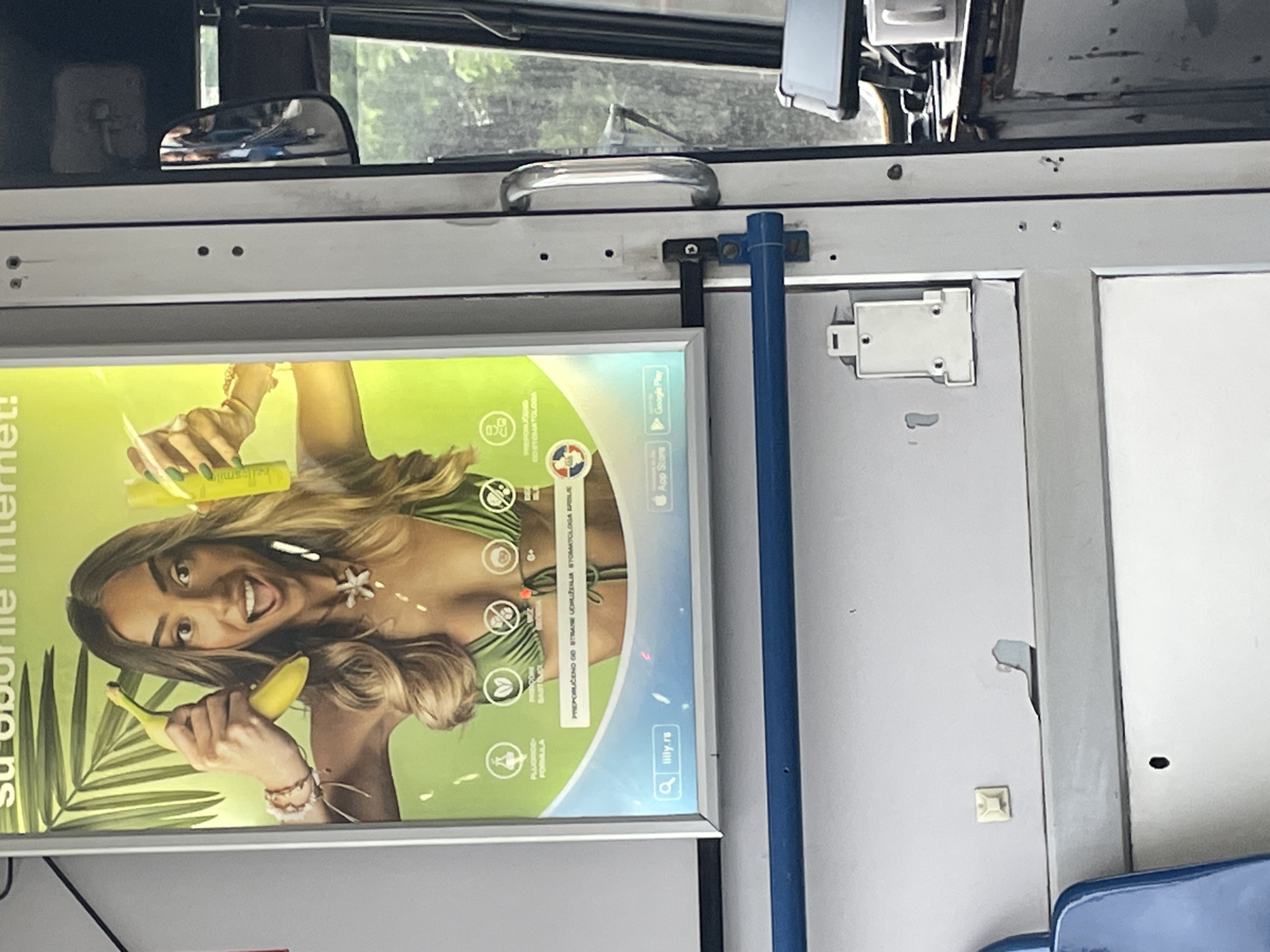 The image shows the interior of what appears to be a public transportation vehicle, possibly a bus or tram, in Belgrade, Serbia. The view is directed at the roof area of the vehicle, featuring a safety handrail, an advertisement display, and various fixtures.
The advertisement display, prominently positioned, features a woman holding a banana and what appears to be a teeth whitening product. The ad includes the text "su oborite Internet!" and logos for the Apple App Store and Google Play Store, along with a website address "illy.rs." The ad also contains text in Serbian: "PREPORUČENO OD STRANE UDRUŽENJA STOMATOLOGA SRBIJE" (Recommended by the Association of Stomatologists of Serbia) and other details.
The vehicle's interior is light gray, with hints of a window and objects outside visible in the upper part of the frame. There are also traces of damage, such as peeling paint, on the interior surface. A blue handrail is situated next to the advertisement display. The photo has been taken at an angle, resulting in a slightly skewed perspective.